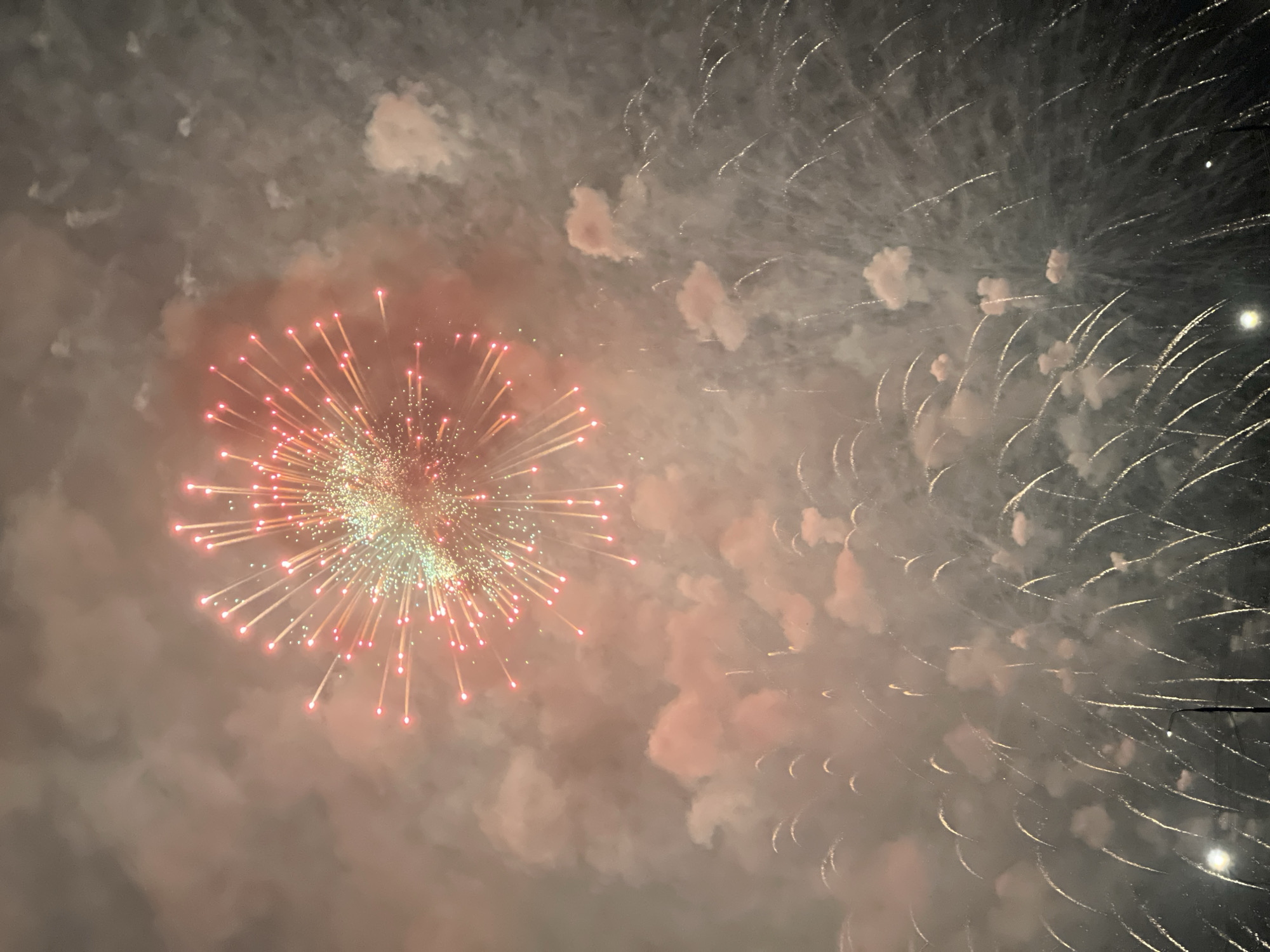 The image depicts a fireworks display, with a central burst of light emanating outwards in a radial pattern, featuring red-tipped and green-centered sparks. To the right, there are arcs of light trails, suggesting a different type of firework or the after-effect of a previous one. The scene is filled with smoke or haze, possibly from the fireworks themselves, which obscures the background. Based on the presence of fireworks, it is likely a celebration. The location is unknown, and there is no visible evidence to suggest the city of Krasnoyarsk, Russia. No people or objects other than the fireworks are visible.