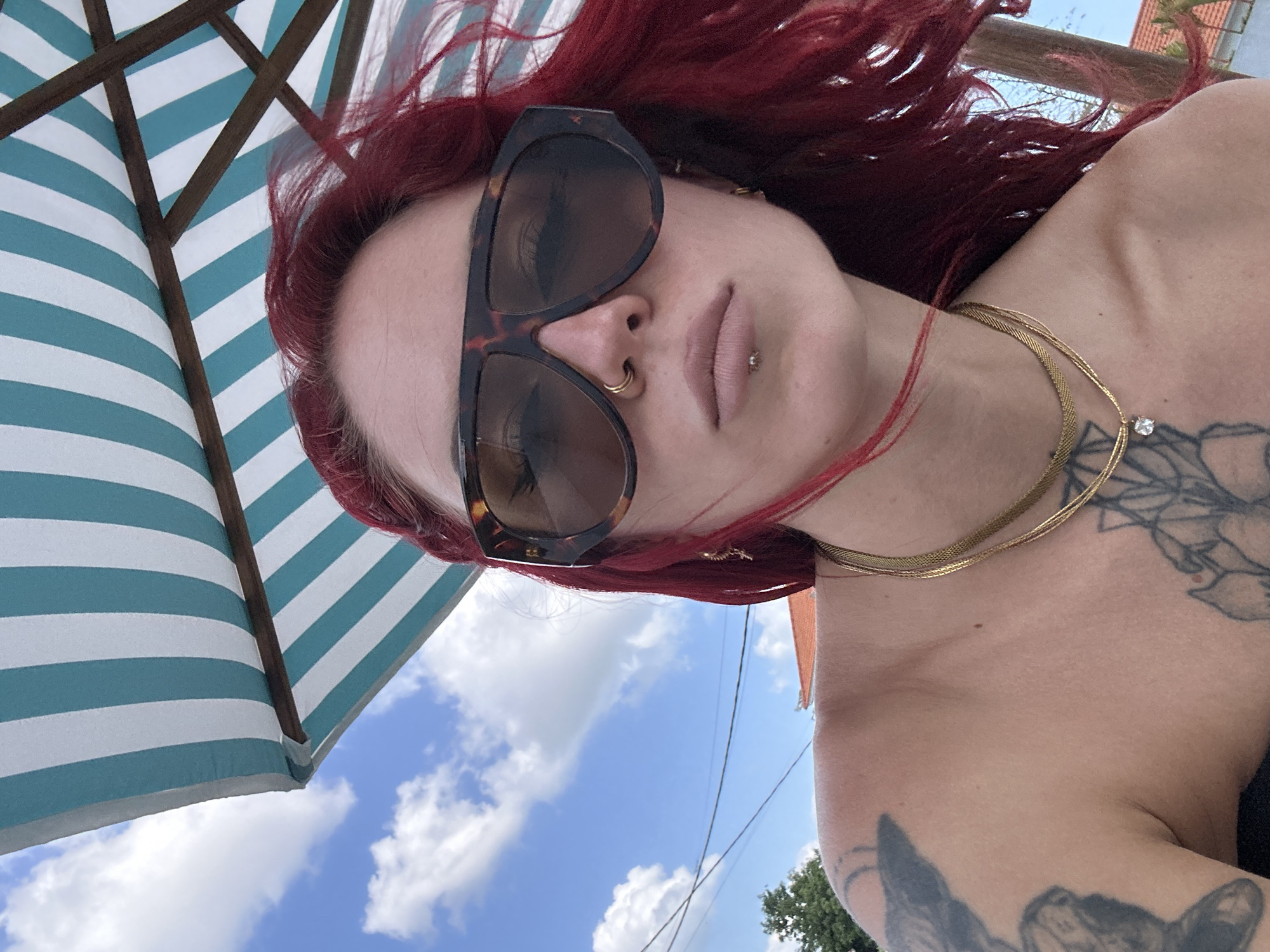 A woman with bright red hair, wearing large tortoiseshell sunglasses, a nose ring, and a lip ring, is pictured outdoors, possibly at a beach resort or a waterfront location in North Kuta, Indonesia. She is leaning back slightly, with her shoulders bare. The background shows a blue sky with white clouds, and a striped umbrella with blue and white stripes is visible overhead, providing shade. Part of a building with an orange roof can be seen in the distance, along with some greenery. The woman has tattoos on her left arm and chest, and she is wearing two gold necklaces. The overall impression is a casual, sunny day.
