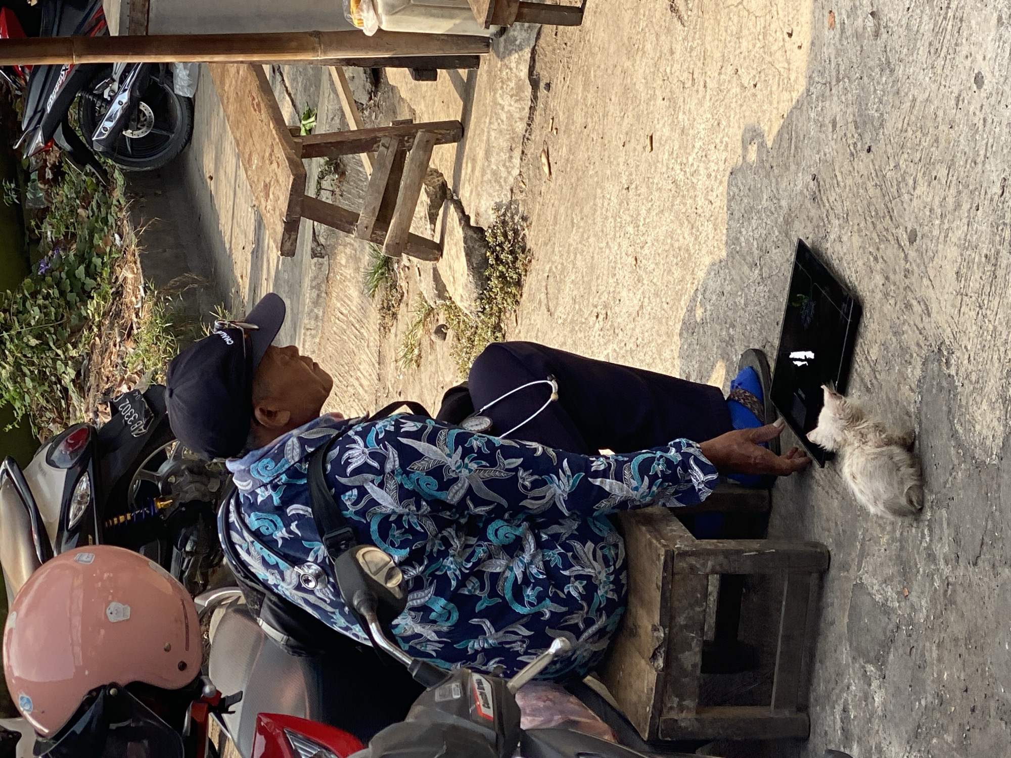In Ambarketawang, Indonesia, a man sits on a wooden stool next to a motorcycle. He wears a dark baseball cap, sunglasses perched on top, and a dark blue patterned shirt. In front of him, a small, light-colored cat sits on the concrete ground, looking at a black tablet. The environment is outdoors, with a cobblestone-like pathway and a wooden bench visible in the background, suggesting a street or market area.