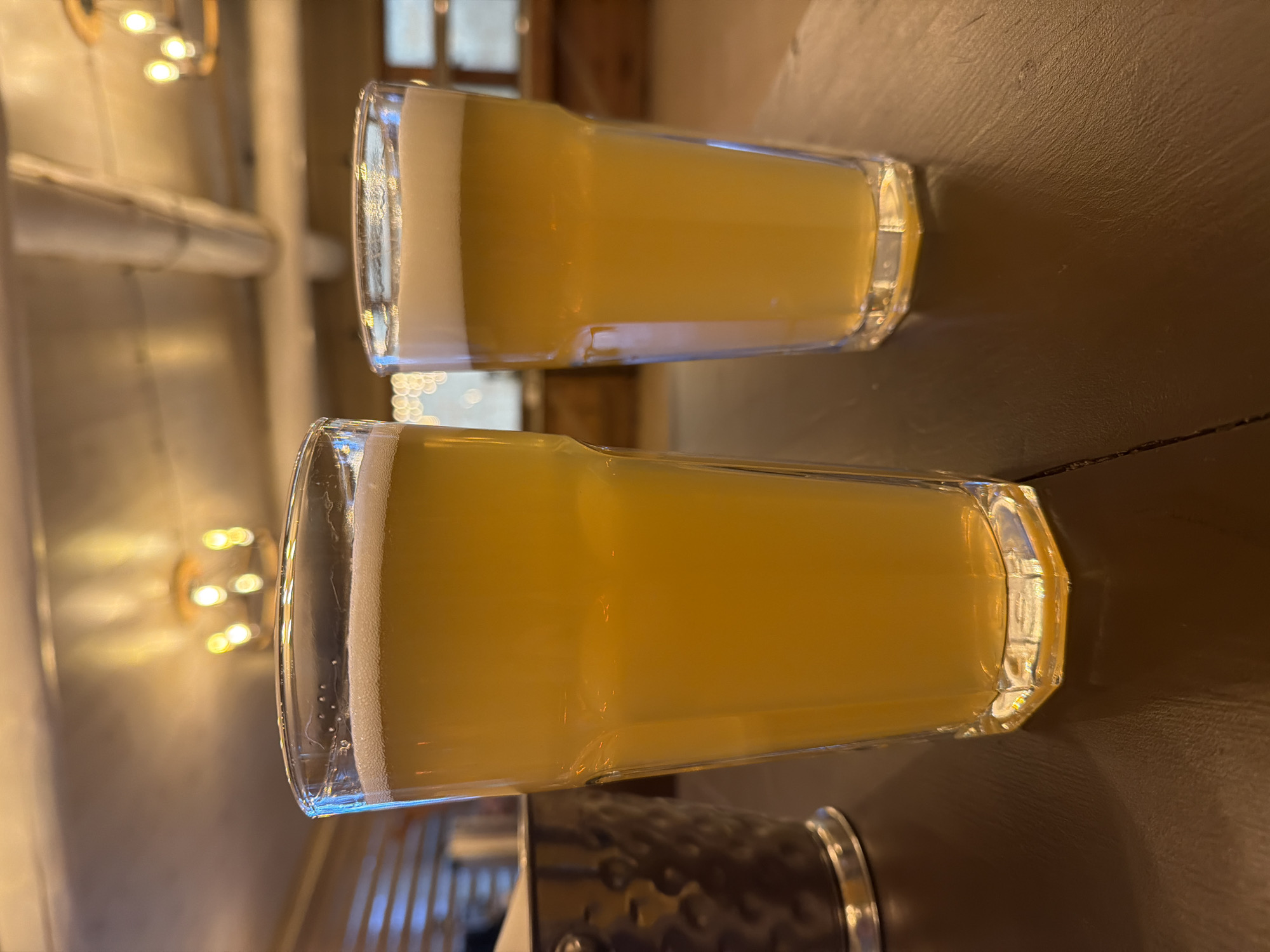 Two glasses of beer with white foamy heads are sitting on a dark, possibly wooden, surface. The beer appears to be a light color, possibly a pale ale or IPA. The glasses are octagonal in shape. In the background, there is a wall with lights and what appears to be a window or opening. The location is not evident from the image. The city of Zaplav'e, Russia is not discernible. There is no information on people, subjects, actions, or interactions visible in the image.