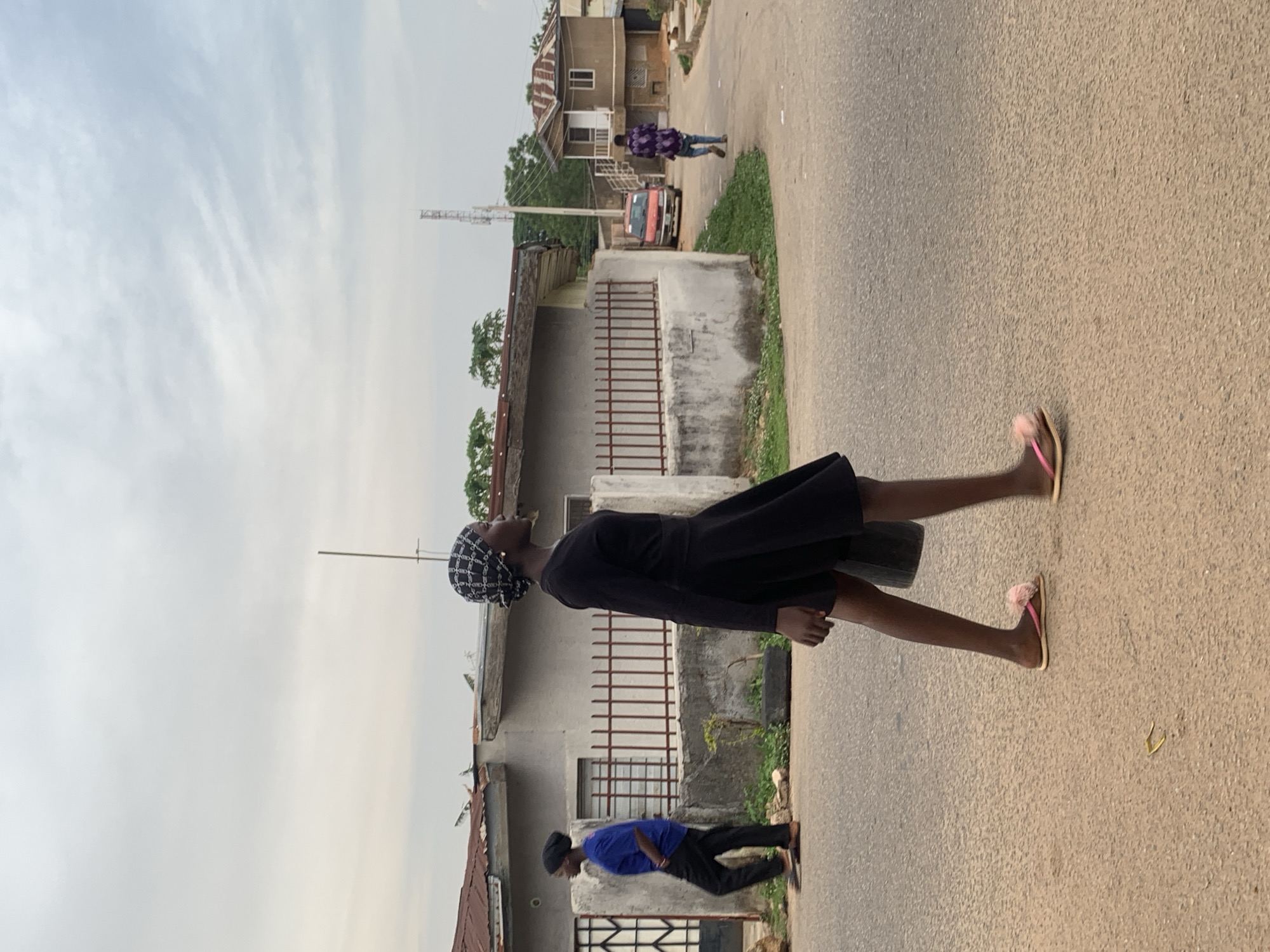 A vertical shot depicts a young woman in a black dress and patterned head covering walking on a paved road in Jos, Nigeria. She is facing toward the sky. The road runs between buildings, some with fenced areas. In the background, a taller communications tower rises above the rooftops. Two figures, one in blue, walk on the road in the same direction, and another is seen further away, walking in the opposite direction. The sky is overcast and light.