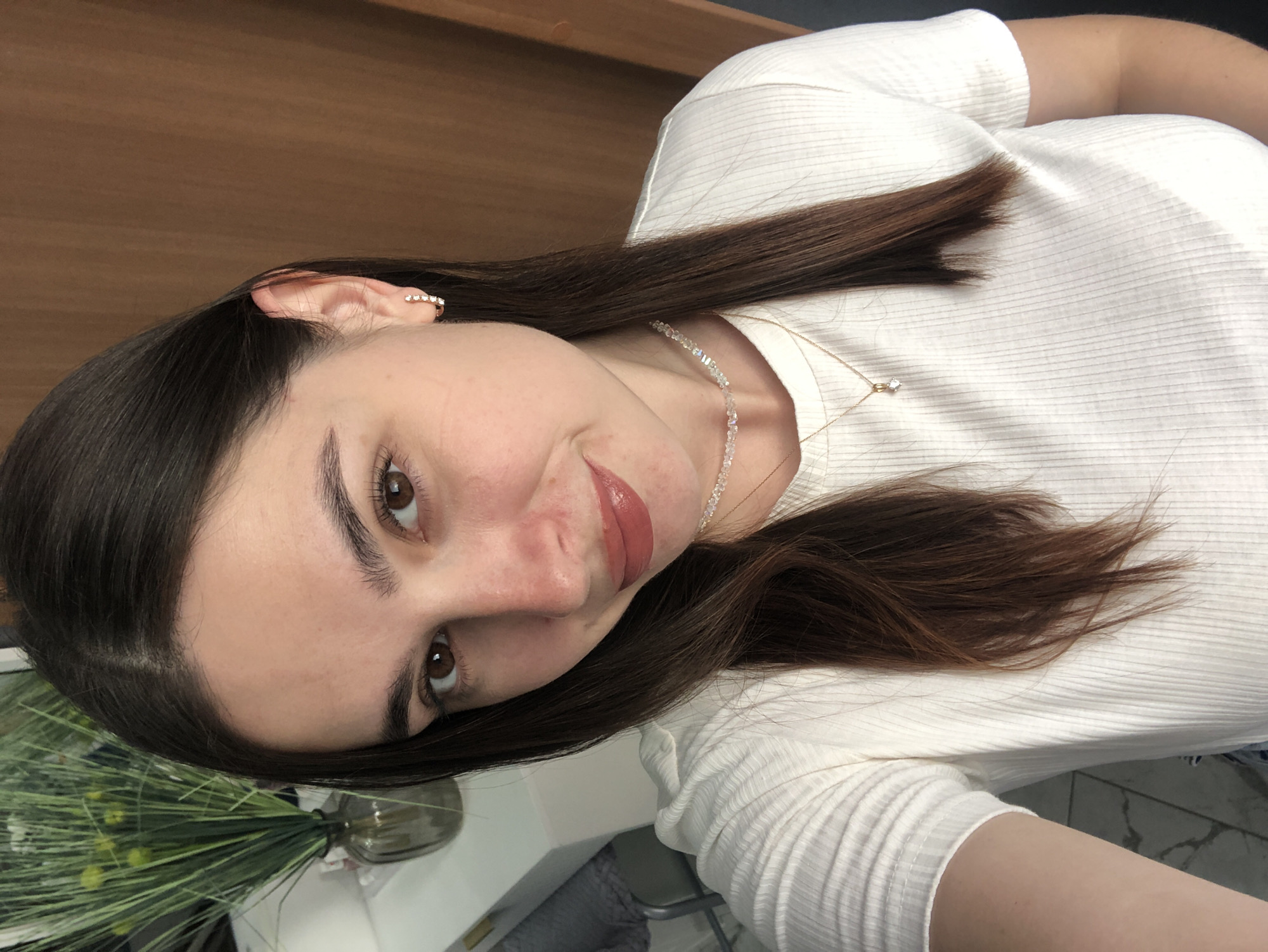 This is a close-up, rotated selfie of a young woman with dark brown hair and brown eyes. She is wearing a white, ribbed t-shirt and a delicate necklace with a small diamond pendant. A small hoop earring with three small diamonds is visible on her left ear. Her makeup includes mascara and lip gloss in a rosy-brown shade. She has a neutral expression, with a slight smile. The background appears to be indoors, with a wooden wall or furniture to the left and what looks like a plant in a vase on a white surface to the lower left. The lighting suggests it could be daytime, possibly indoors with artificial or natural light. No text is visible in the image. The context provided states the location as Chaikovsky, Russia.