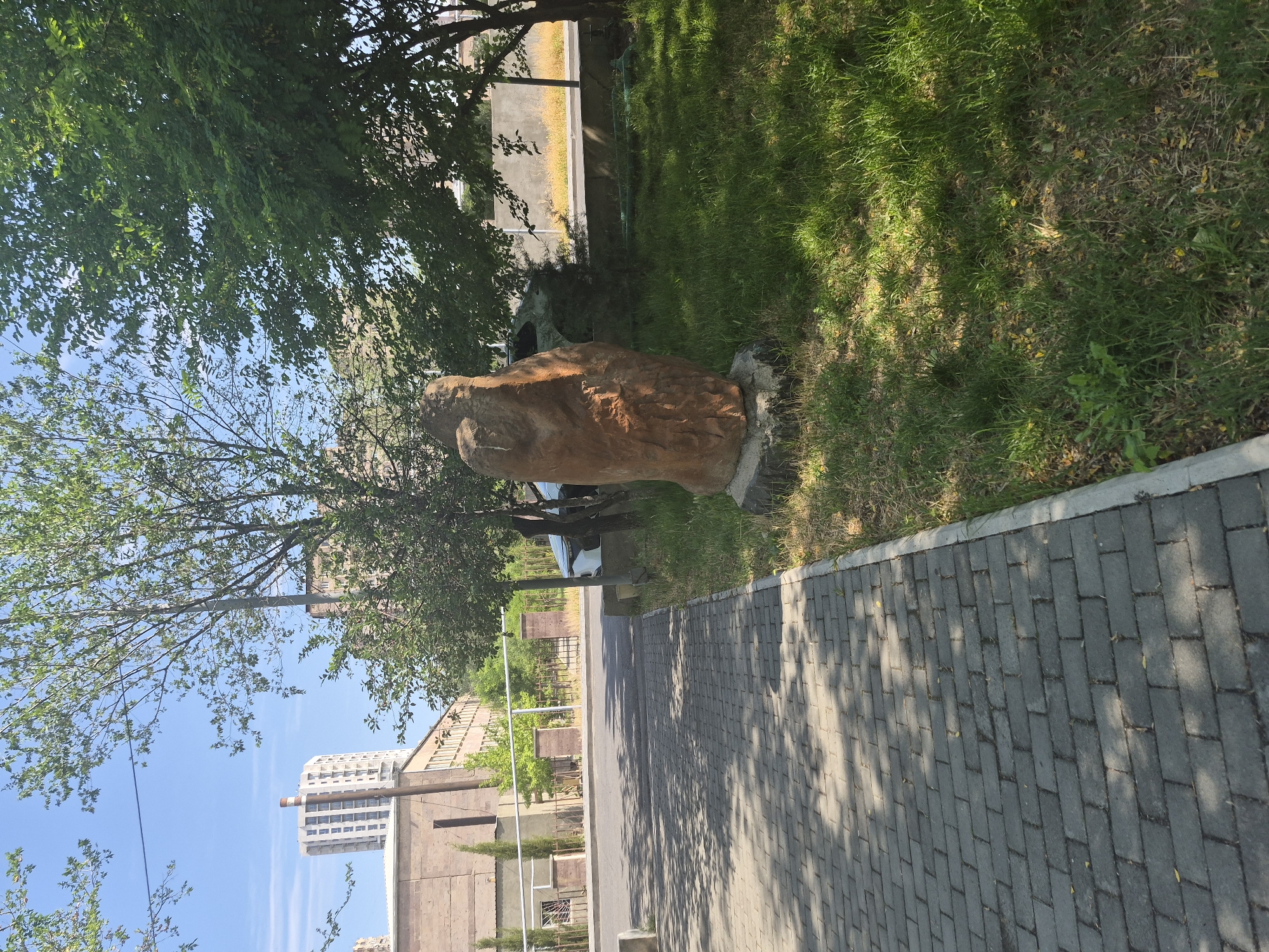 The image captures a street-level view in Yerevan, Armenia, on a bright, sunny day. In the foreground, a gray brick walkway lines the left side of the frame, casting dappled shadows from overhead trees. To the right of the walkway is a grassy area with a tall, reddish-brown stone sculpture positioned on a concrete base. Beyond the sculpture, lush green grass covers the ground, and several trees provide shade.
In the middle ground, a paved road runs parallel to the walkway. A white car is parked near the right edge of the road, partially obscured by trees. Behind the road, a low fence and a wall delineate the property line. In the background, there's a long, light-brown building with multiple windows. Further back, a tall, modern building with a light-colored facade and a prominent vertical pipe on its roof stands against a clear blue sky. The overall scene suggests a well-maintained urban environment with elements of both nature and architecture.