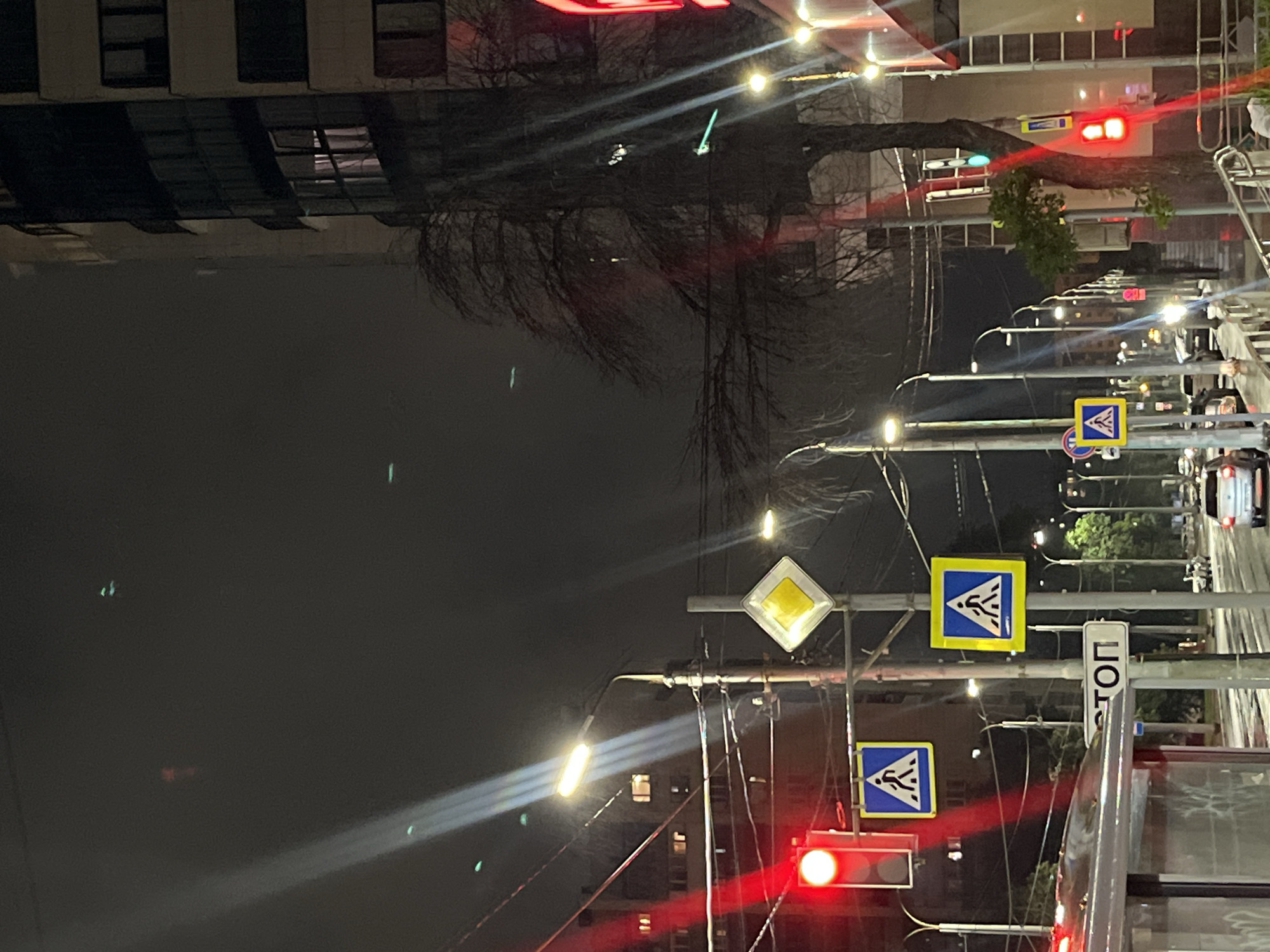 The image depicts a street scene at night, likely in Novokuybyshevsk, Russia. The perspective is from street level, looking up and across the road. The dominant subjects are street signs, traffic lights, and a leafless tree. A traffic light is visible, showing a red light, and another traffic light with a green light is also visible in the background. There are multiple pedestrian crossing signs, which are square with a blue background and a white triangle containing a silhouette of people crossing. A diamond-shaped sign with a yellow background, often indicating a main road, is also present. A vertical sign with Cyrillic text "СТОП" (STOP) is visible.
Streetlights illuminate the scene, casting long beams of light, suggesting a clear but dark night. The leafless branches of a tree extend across the upper portion of the image, silhouetted against the ambient light and the buildings in the background. The buildings themselves appear to be multi-story structures with windows. Vehicles are present on the road, indicated by headlights and taillights, suggesting traffic flow. One car is clearly visible in the background. The overall atmosphere is urban and functional, with the emphasis on traffic control and pedestrian safety. There are no visible people in the image.