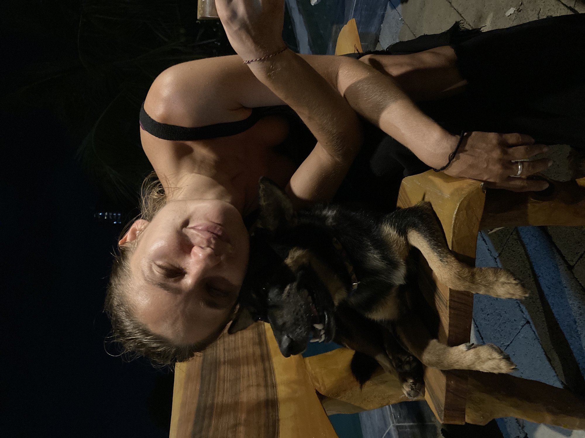 A woman with fair skin and wet hair is shown indoors at night in Mũi Né, Vietnam. She is wearing a black top and is seated in a wooden chair. She has a ring on her finger. A German Shepherd dog rests in her lap. The dog is mostly black with tan markings on its legs. The chair is positioned on a blue tiled surface.