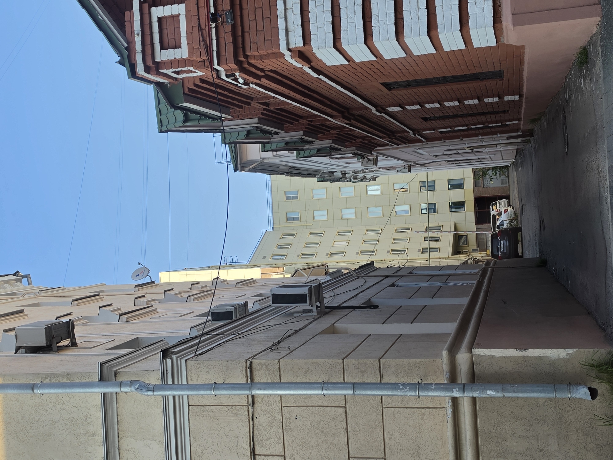 The image depicts a narrow alleyway between two buildings. On the left is a light-colored building with architectural detailing and visible air conditioning units. A grey downspout runs vertically along this building. On the right is a red brick building with white decorative elements and a green roof. Further down the alley, a pale yellow, modern apartment building is visible. Two cars are parked in the distance. The sky is clear and blue. The image appears to be taken in Nizhny Novgorod, Russia, given the architectural styles and general urban setting.