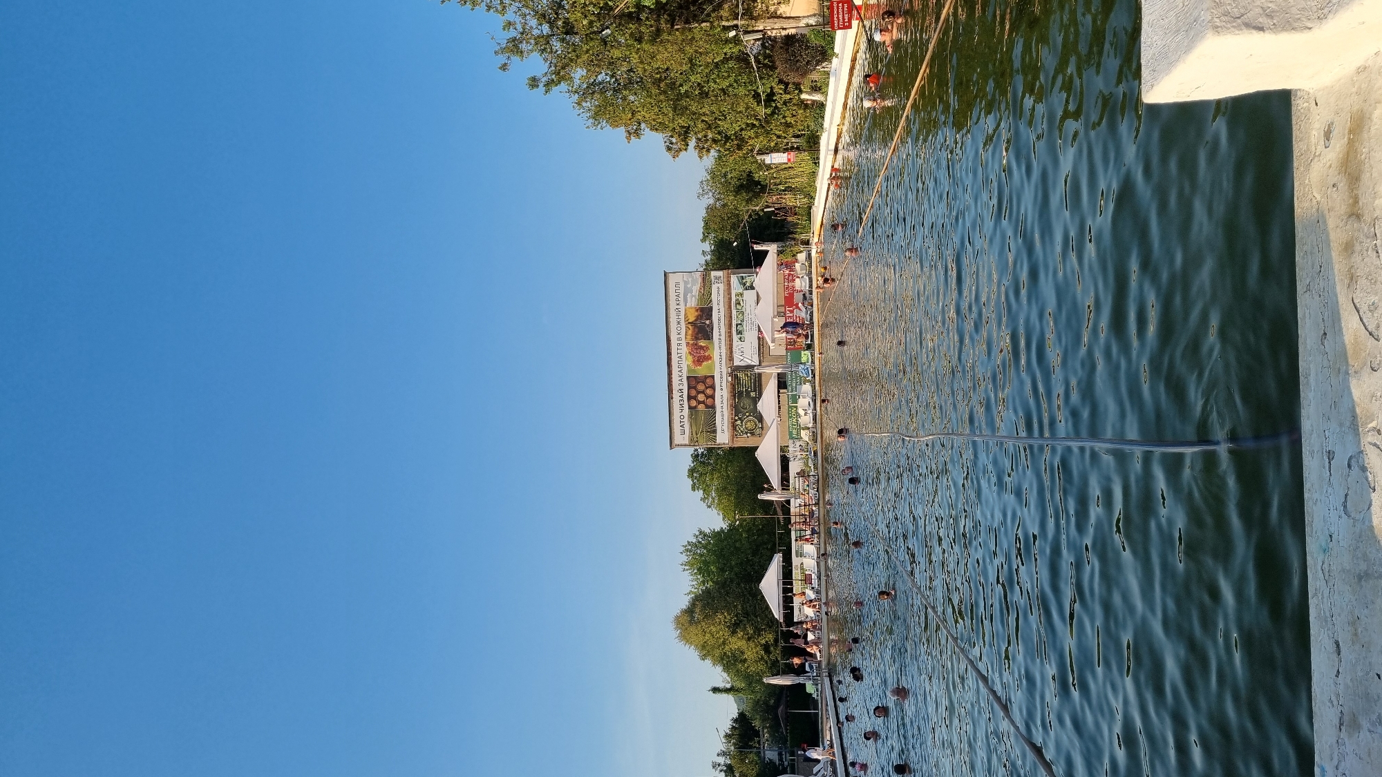 The image shows an outdoor swimming pool filled with people swimming and lounging. The pool is rectangular and surrounded by a white concrete edge. In the background, there are several white gazebos with lounge chairs, trees, and a large billboard advertising "ШАТО ЧИЗАЙ ЗАКАРПАТТЯ В КОЖНІЙ КРАПЛІ," which translates to "Chizay Castle Transcarpathia in Every Drop." This billboard also features images of grapes and wine. The location appears to be a resort or recreational area in Berehove, Ukraine, known for its thermal waters and wineries. The sky is clear and blue, indicating a sunny day. A rope stretches across the pool, possibly for lane separation or safety. A red and white sign with Cyrillic text is visible on the right side of the image, near the edge of the pool.