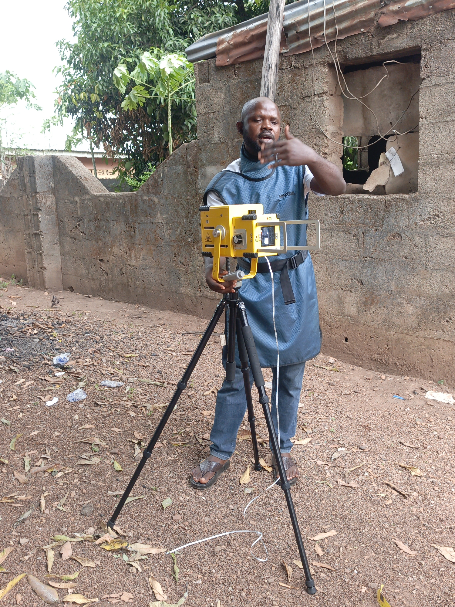 A man stands outdoors in Akwanga, Nigeria. He is setting up a yellow radiation measurement device mounted on a black tripod. The man wears jeans, sandals, a white t-shirt, and a blue apron. He is gesturing with his right hand. Behind him is a partially constructed brick wall with a rectangular opening. A corrugated metal roof is visible on the wall. The ground is dirt and gravel strewn with leaves and trash. A tree is visible in the background.