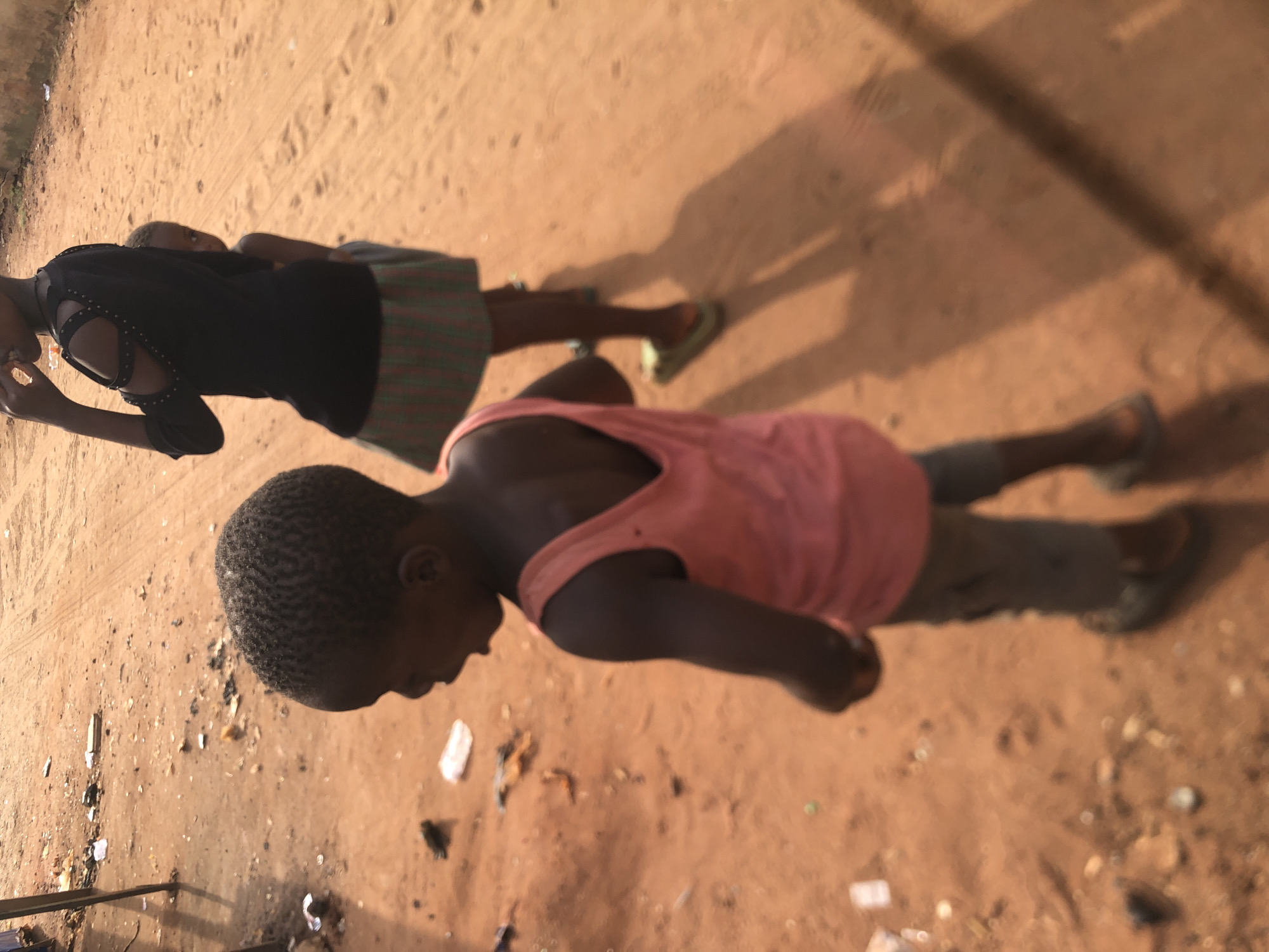 The image, taken with a vertical orientation (rotated 90 degrees clockwise), captures two children on a dusty, light brown ground in an unpaved outdoor setting, likely in Onitsha, Nigeria.
In the foreground, a child with short, dark, curly hair is visible from the waist up, wearing a pink sleeveless tank top and dark pants or shorts. Their face is partly obscured as they look downwards. Their arms are relaxed at their sides.
Behind and slightly to the right, another child stands, partly out of focus. This child is wearing a dark top with decorative cut-outs on the shoulders and a horizontally striped skirt in shades of green and a darker color. They are wearing light green flip-flops on their feet. Their facial features are not clearly discernible.
The ground is a consistent light brown, appearing sandy or dusty, and is littered with small pieces of debris, including white paper fragments and various dark speckles. Long, sharp shadows of people are cast prominently across the ground, indicating strong, low-angle sunlight, typical of either morning or late afternoon. The scene conveys a casual, everyday outdoor moment under a clear, sunny sky. There is no visible text in the image.