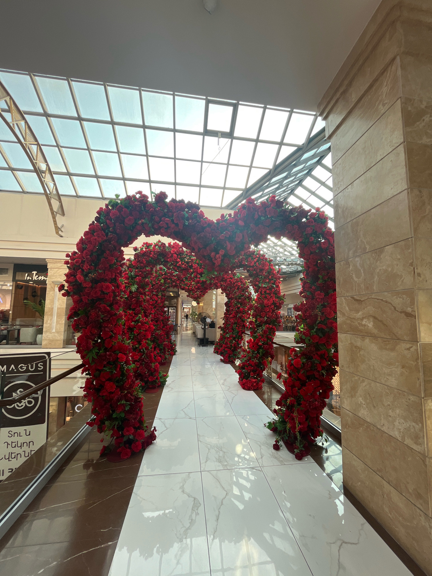The image captures a decorative floral archway made of red roses, forming a heart shape, in what appears to be an indoor public space. The archway is centrally positioned, leading into a walkway. The walkway is paved with white and brown marbled tiles, reflecting the overhead lighting. Large windows or a skylight structure dominate the upper portion of the image, allowing natural light to filter in. The surrounding architecture features beige marble-clad columns and walls. Signs with Armenian text are visible on a railing to the left, one of which reads "MAGUS". The setting suggests a shopping mall or a similar commercial or event venue. A few individuals are visible in the background, walking through the space, but they are not the main focus. The overall ambiance is festive and decorative, likely for an event or seasonal display. Based on the "MAGUS" sign which is a prominent shopping center in Yerevan, Armenia, this scene is likely located in the MAGUS Mall in Yerevan.