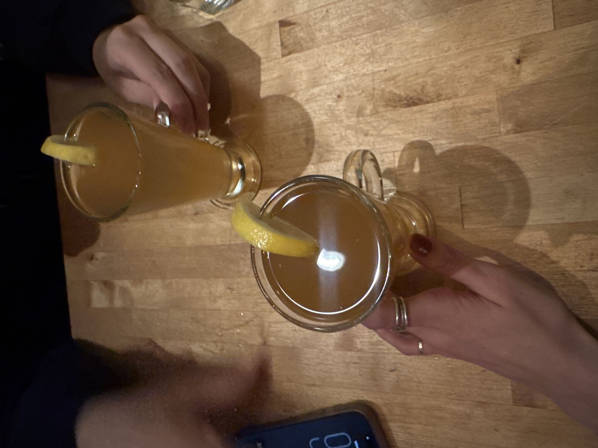 The image shows two glasses of amber-colored liquid, likely beverages, placed on a wooden surface. Each glass is garnished with a slice of lemon. Two individuals are partially visible in the frame. One person, on the left, is holding a glass. The other person, on the right, is also holding a glass and has visible rings on their fingers. A smartphone is placed near the lower part of the frame. The wooden surface appears to be a table. It is not possible to determine the location as New York City.