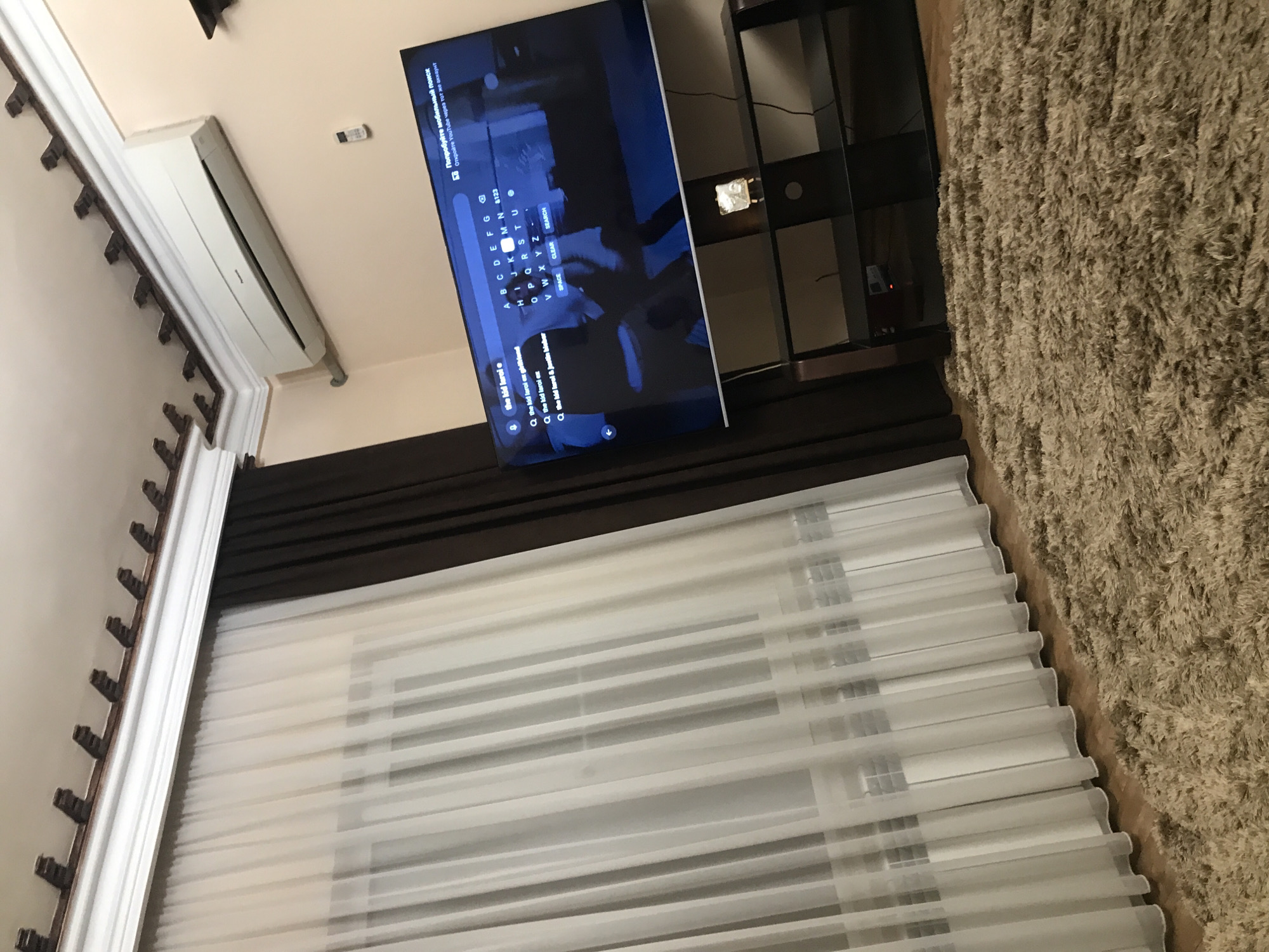 The image shows an indoor scene. A television displays a search interface with the phrase "the kid laroi e" and related suggestions. A media stand sits next to the TV. The bottom of the image is a window with blinds and dark curtains. A beige rug is on the right side of the image. There is also an air conditioning unit mounted high on the wall. Based on the OCR text, the location may be Yerevan, Armenia.