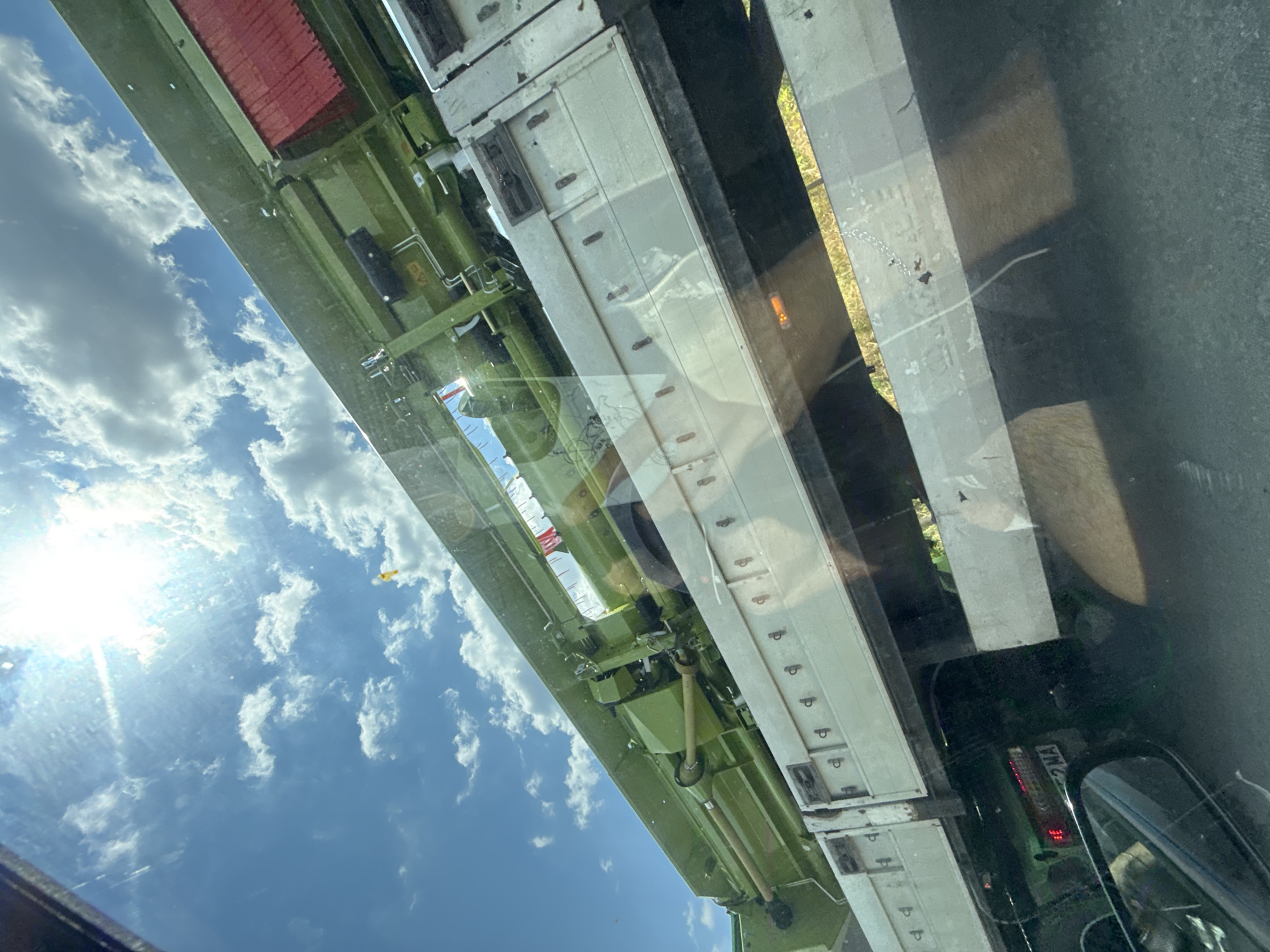 This media file captures a reflection in what appears to be a vehicle's window, oriented sideways. The reflection shows a bright, sunny sky filled with fluffy white clouds, indicating a clear day. The sun is a strong, bright source of light, creating lens flare and highlighting the texture of the clouds.
The main subject of the reflection is the side of a large, industrial vehicle, likely a truck or trailer. It has a long, segmented body, primarily painted in a pale, off-white color. Below this main body, there is a complex green metal structure, possibly part of the vehicle's undercarriage or a piece of machinery. This green section is detailed with various pipes, hoses, and mechanical components.
In the distance, through the reflection, a hint of a landscape with greenery and sunlight can be seen. The foreground of the reflection shows a dark, possibly road surface, with reflections of streetlights or vehicle lights visible as orange and red glows.
There are some notable details within the reflection: a small yellow object, possibly a flag or marker, is visible against the blue sky. On the white segmented part of the vehicle, there are regularly spaced dark markings, possibly hinges or latches. The overall impression is of a daytime scene, likely afternoon given the bright sunlight. The perspective suggests the viewer is inside another vehicle, looking out and seeing the reflection of the sky and the vehicle beside them. No people are clearly discernible in the reflection. There is a license plate visible on a vehicle in the distance within the reflection that reads "2 MA".