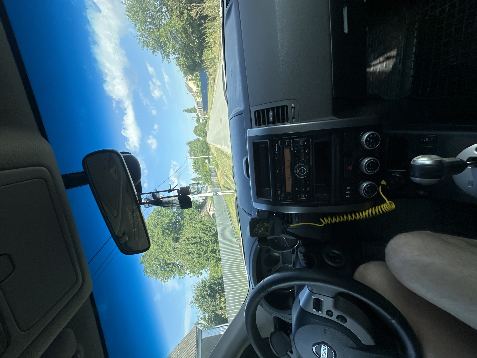 The image shows the interior of a car, looking forward through the windshield. The view is of a paved road in a rural or suburban area, with houses and trees visible on either side. The sky is blue with some scattered clouds. The car's dashboard is visible in the foreground, with a steering wheel on the left and a car stereo system in the center. The driver's legs are also visible. The location is identified as Makiv, Ukraine.