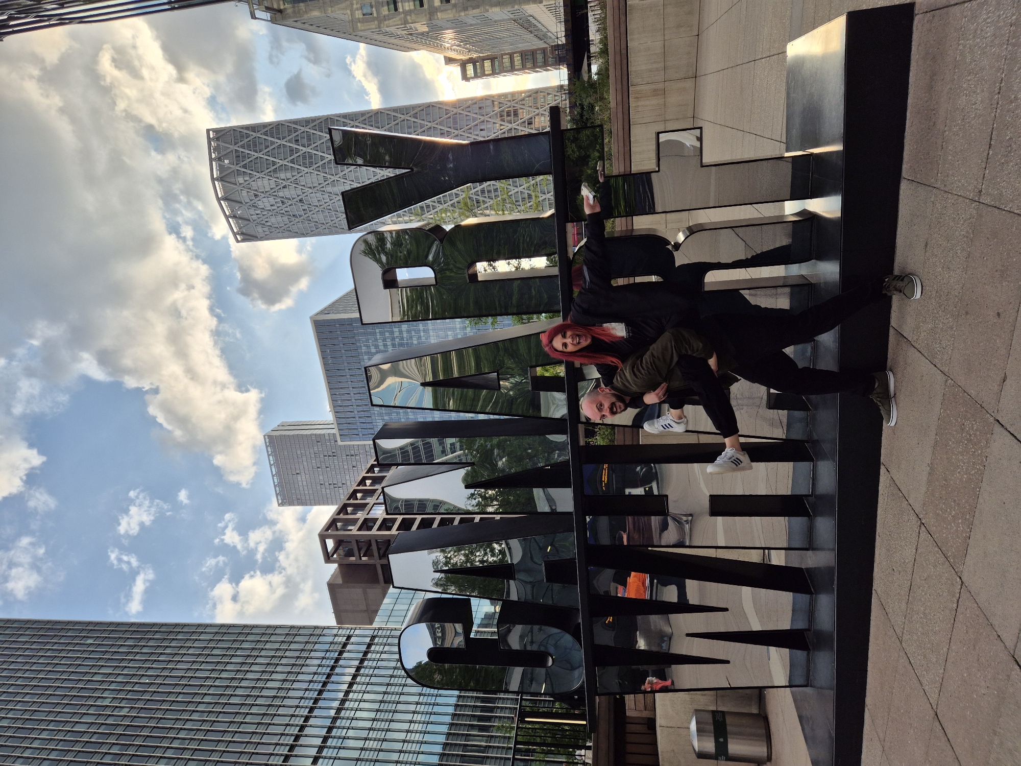 The image shows a man and a woman in front of a large metal sign that spells out "CANADA SQUARE". The man is wearing a green jacket and white sneakers, while the woman has red hair and is wearing a black jacket. They appear to be interacting with the sign, possibly posing for a photo.
The location is Canada Square, in the Canary Wharf area of London, United Kingdom. Modern office buildings and a blue sky with clouds are visible in the background. The sign is placed on a stone paved area.