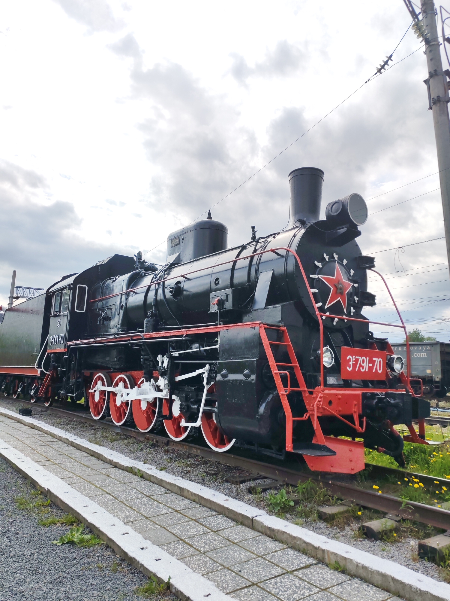 This image features a large, black Soviet-era steam locomotive, identified as model ЭМ 791-70, on static display outdoors in Medvezhyegorsk, Russia. The locomotive is the main subject, presented from a low-angle view that highlights its impressive size and detail. Its robust black body is accentuated by vibrant red elements, including the large driving wheels, the front bumper, and various handrails and ladders. A prominent red five-pointed star is affixed to the front of the boiler.
The locomotive bears the identification number "3*791-70" in white text on a red plate at its front, and "ЭМ791-70" is also visible on the side of its attached tender car. It rests on a set of railroad tracks, alongside which a paved walkway of square grey tiles is laid out, suggesting a dedicated viewing area for visitors. No people are visible in the scene.
The setting is an open-air environment under an overcast sky with scattered clouds, indicating it is daytime with subdued, cloudy weather. In the background, on the right side, a utility pole with electrical wires extends upwards. Further down the tracks, a glimpse of another train car, possibly a freight wagon, is visible, with faint text that appears to be "truzovaya.com". The image captures a moment of historical preservation, showcasing a significant piece of railway engineering.
