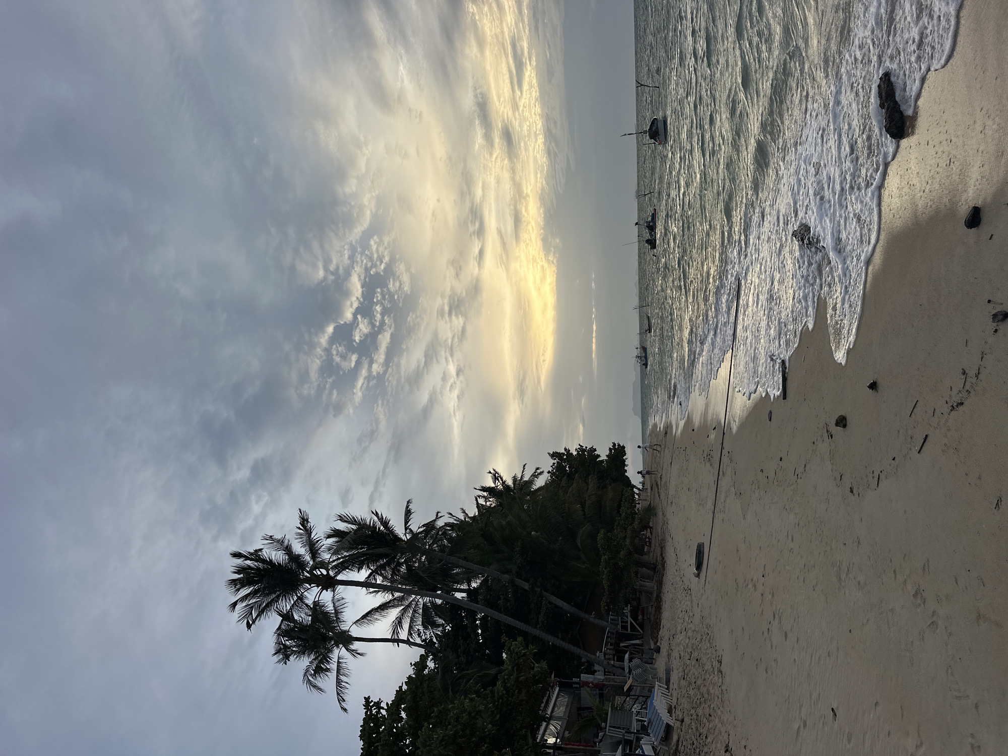 A serene beach scene unfolds in Ko Samui District, Thailand, under a cloudy sky with streaks of golden light breaking through. The sandy shore is lined with lush green palm trees, their fronds silhouetted against the muted sky. The gentle waves of the turquoise sea lap at the shore, creating white foam that washes over the sand. Several small fishing boats are anchored in the distance, their masts visible against the horizon. Two figures can be seen walking along the water's edge, their presence adding a sense of scale and tranquility to the vast expanse of the beach and ocean. The overall atmosphere is one of peacefulness and natural beauty.