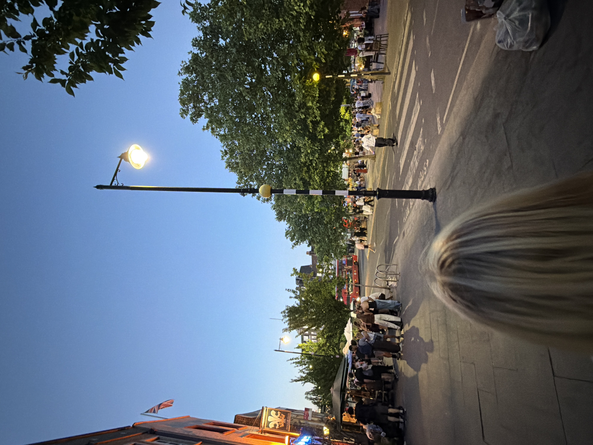 The image captures an outdoor scene at dusk in London, United Kingdom. A street lamp with a lit bulb stands prominently on the left side of the frame. Crowds of people populate the sidewalks and crosswalks. The foreground includes a portion of a person's head with blonde hair. Buildings line the street, some adorned with flags. Outdoor seating areas with tables and chairs are visible. A double-decker bus and pedestrian traffic signal are partially visible in the background. The sky is a gradient of blues, indicating evening light.