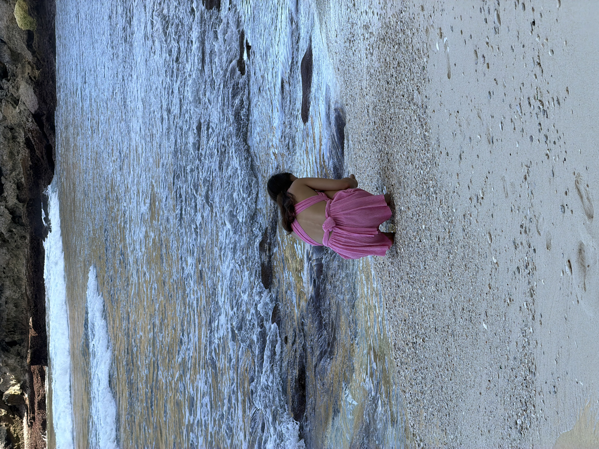 A young girl in a pink dress is crouching on the beach in Pejukutan, Indonesia. She is near the water's edge where the water meets the sand. The beach is composed of a mix of sand and small pebbles. To the left of the image, there is a rocky formation. The overall color palette is a mix of blues, browns, and pinks.