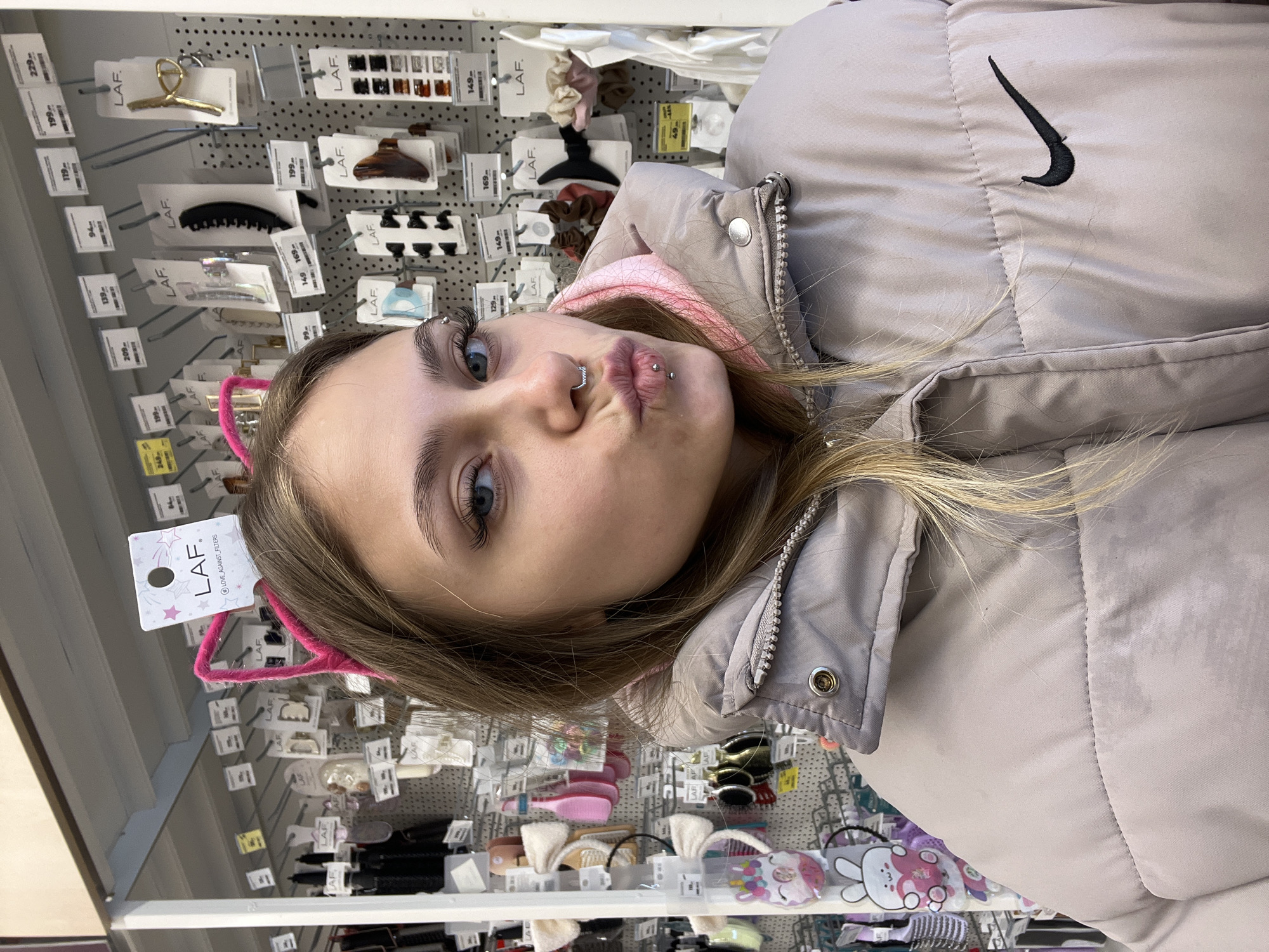 Here is a forensic description of the image:
The image shows a young woman posing for a selfie in what appears to be a retail store. She is wearing a light-colored puffer jacket with a black Nike logo. She also has pink cat-ear hair accessories on. The background consists of a wall display of hair accessories, with labels that include the brand "LAF" and prices. The store is located in the city of Chaikovsky, Russia based on the Cyrillic writing on the price tags. She has multiple facial piercings.