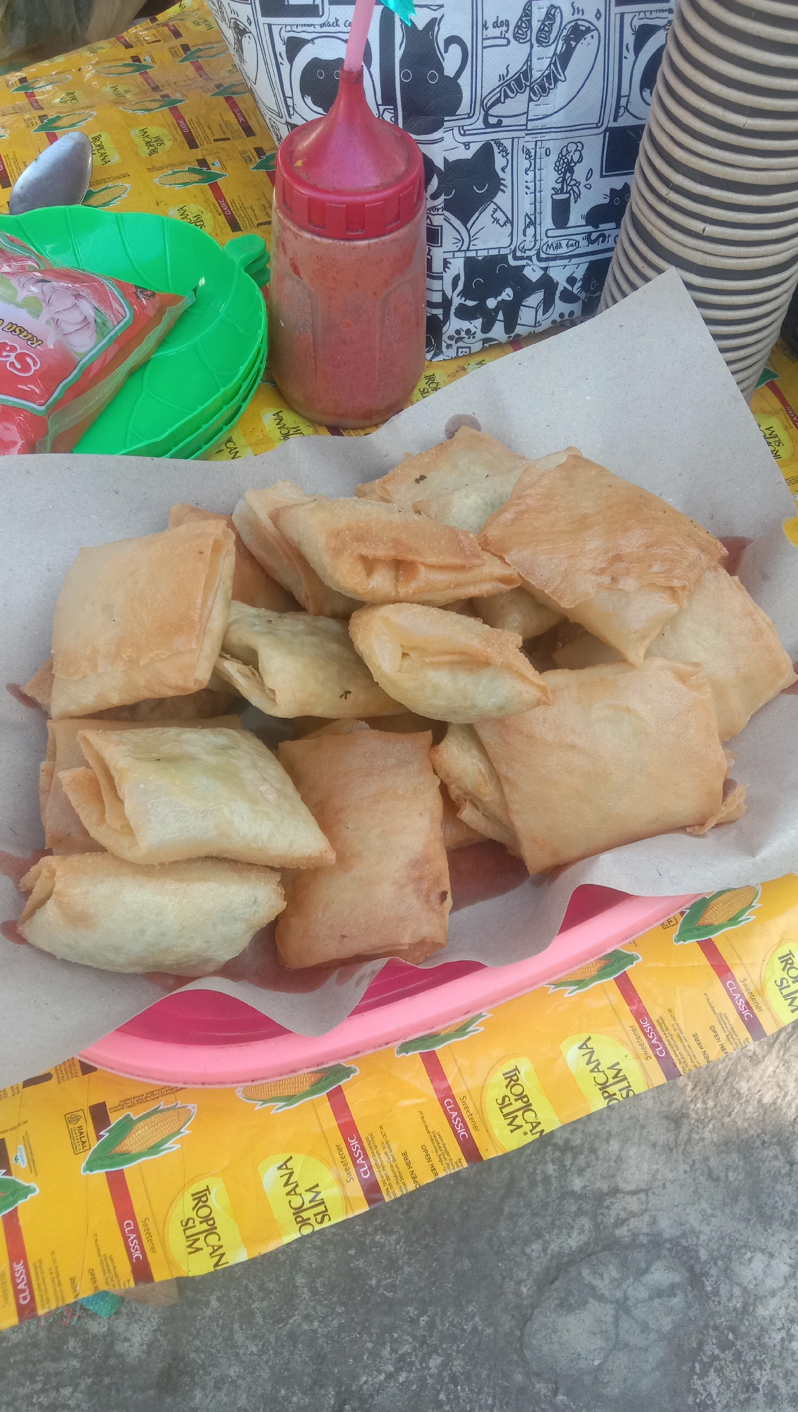A close-up, overhead shot captures a serving of fried snacks, likely lumpia basah, presented in a brown paper wrapper resting on a pink plastic tray. The snacks are golden brown and appear to be filled with a light-colored mixture, possibly vegetables or meat. To the left of the snacks, two green leaf-shaped plastic plates are stacked, with a sachet of sauce peeking out from under the top plate. Behind the plates and snacks, a red squeeze bottle with a red cap, filled with a reddish sauce, stands upright. The background is a yellow cloth with a repeating pattern of corn and text, including "TROPICAN SLIM CLASSIC." A stack of white disposable cups can be seen on the far right. The overall setting suggests an outdoor food stall or casual dining environment in Ngempit, Indonesia. The lighting is bright, indicating daytime.