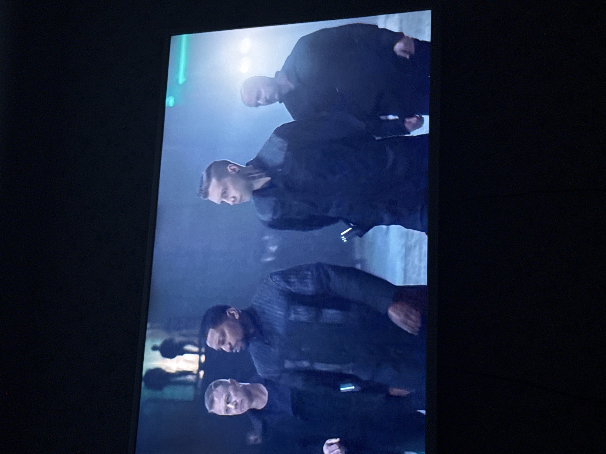 A television screen displays a scene depicting four men in dark clothing. The setting appears to be urban, potentially a street or public space with other people visible in the background, partially obscured by a bluish-grey haze or fog. Illumination comes from a bright light source above. The image appears to be filmed in a street. This image does not display the city of Mytishchi, Russia.