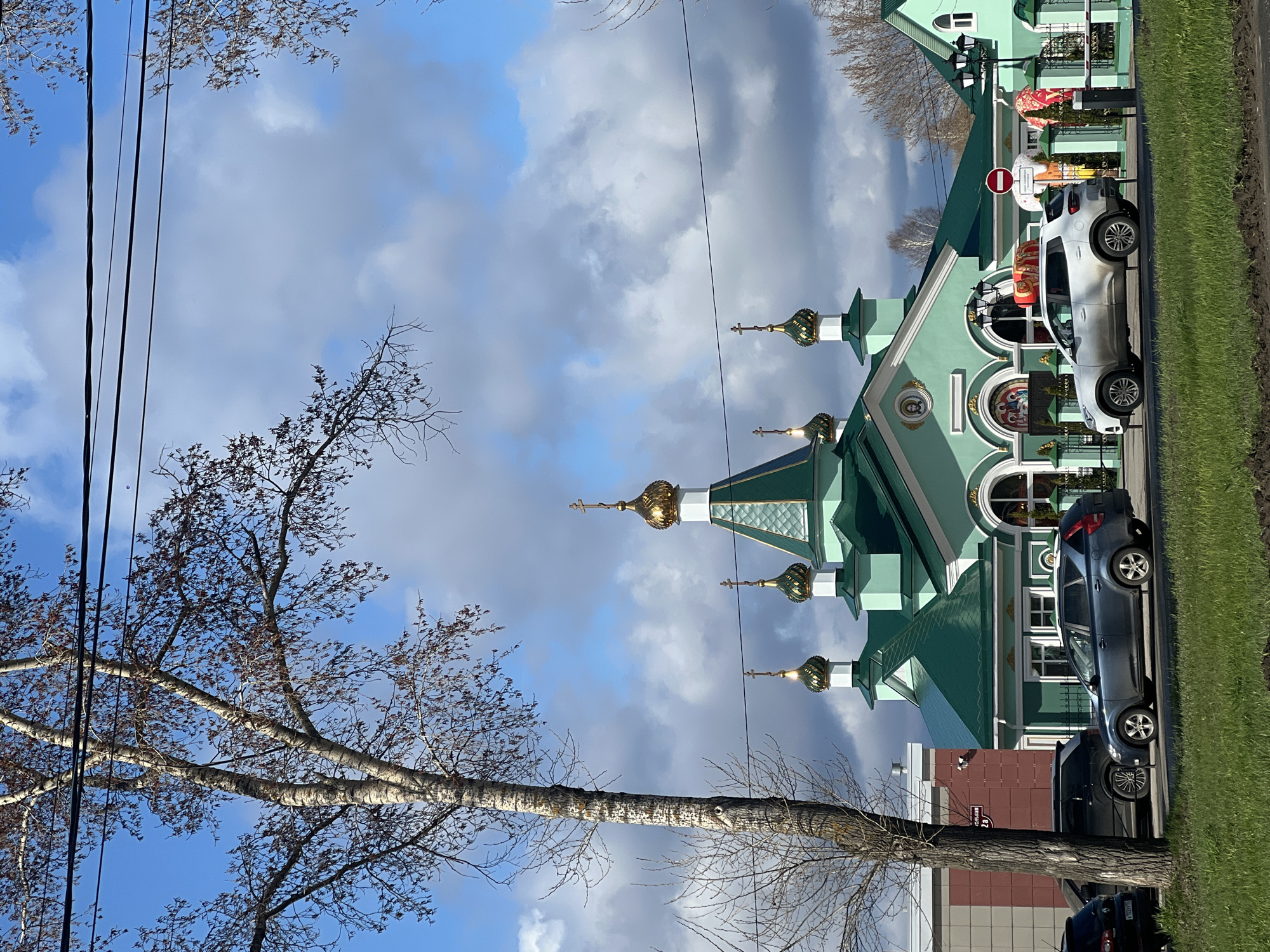 An outdoor scene features a vibrant green-domed church under a partially cloudy blue sky. The church, likely Russian Orthodox, is adorned with multiple golden onion domes, each topped with a cross. Its facade includes arched windows and decorative elements.
In the foreground, a paved road runs parallel to the church, with a strip of green grass separating it from the building. Two cars are parked on the road: a silver/white SUV closer to the church entrance, and a darker grey/black SUV further down. A red circular "No Entry" sign is visible near the front of the silver SUV.
On the left side of the image, a tall, light-barked tree (resembling a birch) with bare branches dominates the foreground, extending upwards and to the right, partially framing the church and the sky. Other leafless tree branches are scattered throughout the upper and left parts of the frame. Overhead utility lines stretch across the sky.
In the background on the right, beyond the church, a building with a reddish-brown brick facade is visible, displaying a street sign with the Cyrillic text "ЧАЙКОВСКОГО 12" (Chaykovskogo 12), indicating a street name and building number.
The scene appears to be set during daytime, with clear lighting suggesting either late morning or afternoon. The bare trees indicate it is likely late autumn, winter, or early spring. No people are visible in the image.