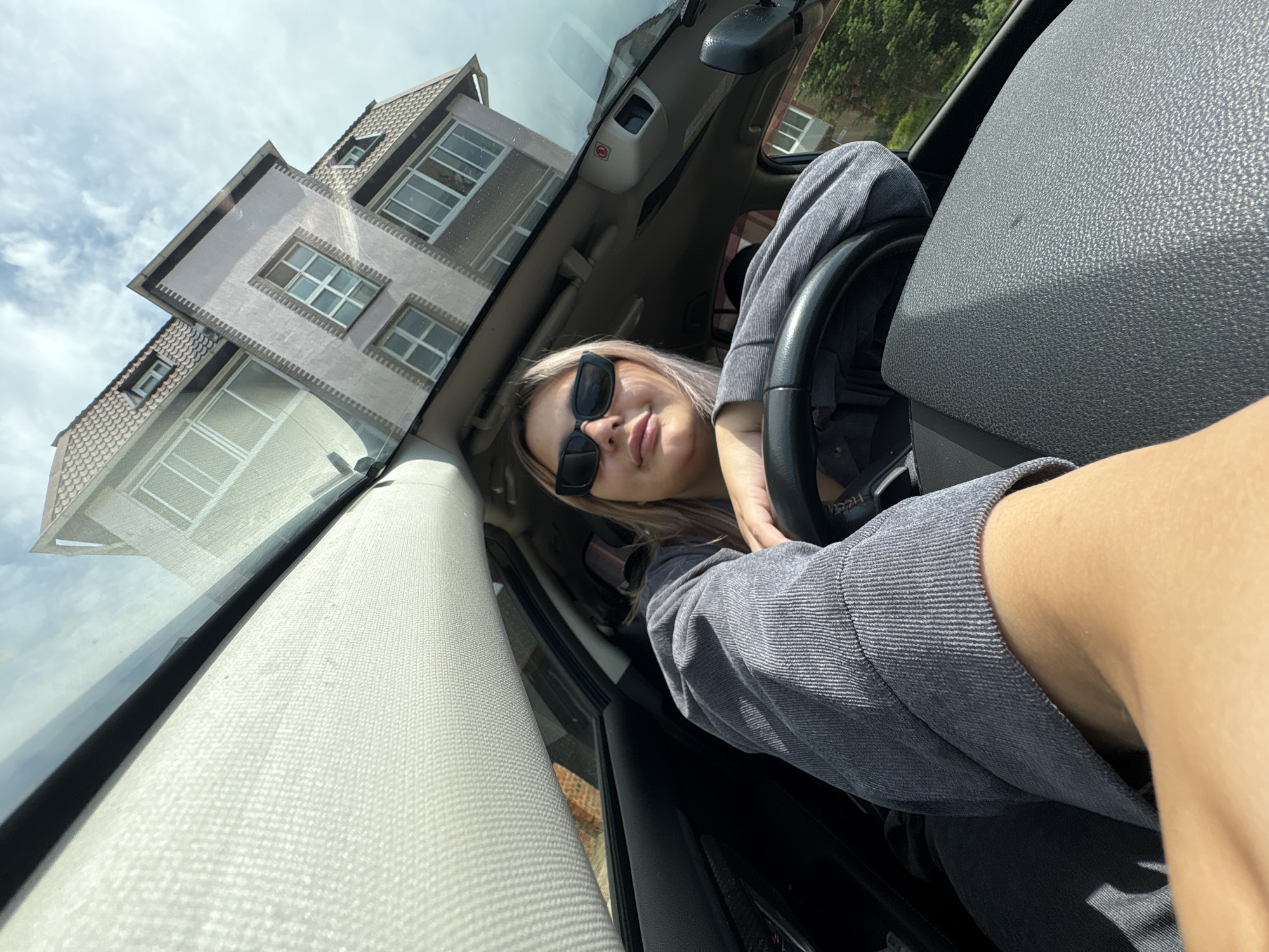 A young woman with blonde hair and sunglasses is sitting in the driver's seat of a car, looking at the camera. She is wearing a gray corduroy jacket and dark pants. The car's interior is visible, including the steering wheel and dashboard. Outside the car, a multi-story building with a distinctive roofline and windows is reflected in the windshield. The sky is cloudy. The scene appears to be a daytime shot, likely in a residential area.
