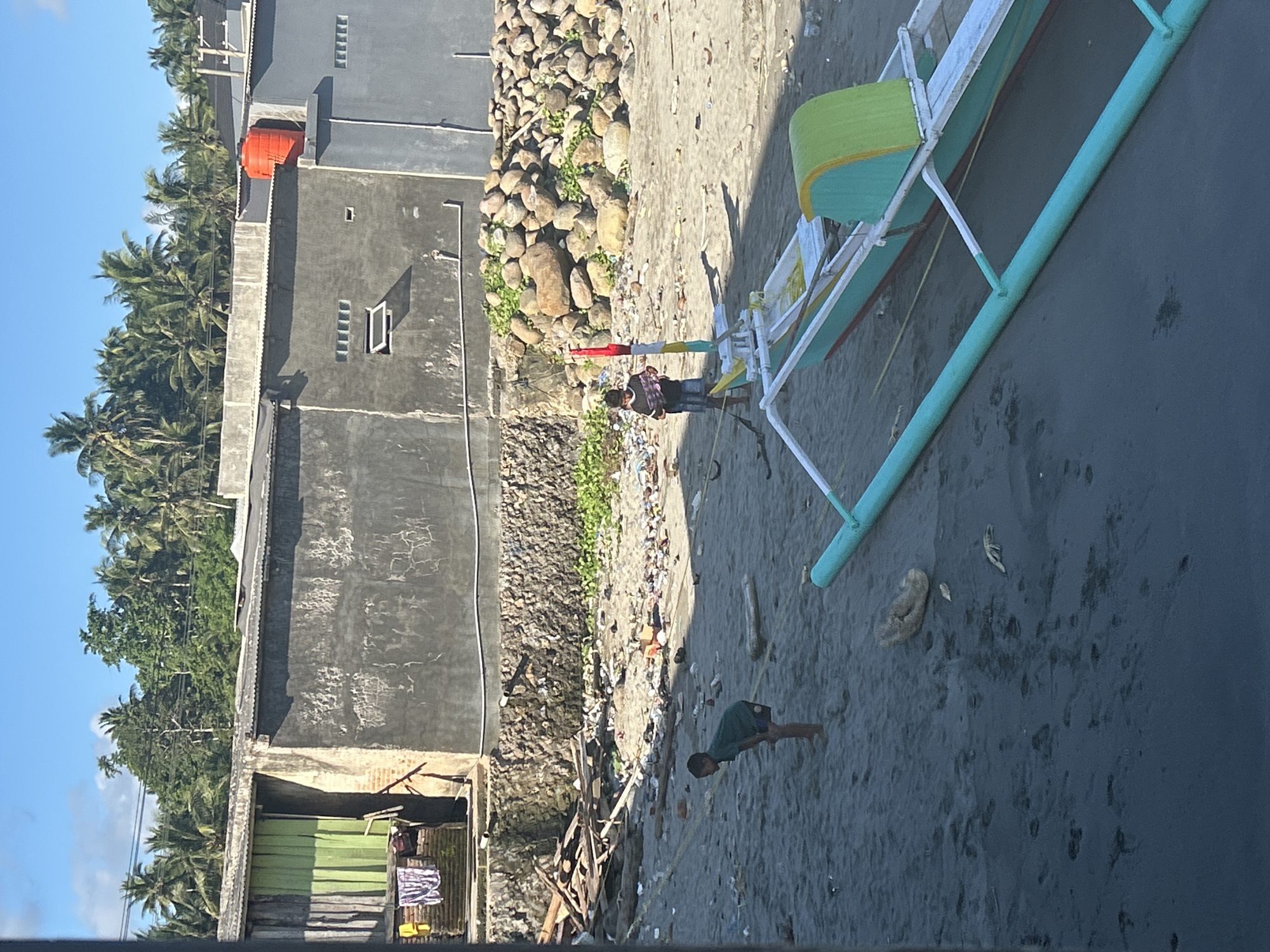 In A R A, Indonesia, a bright, sunny day reveals a beach littered with debris. Two young boys are present on the sandy shore. One boy, wearing a dark green t-shirt and shorts, is crouched near the water. The other boy, in a striped t-shirt and shorts, stands by a colorful outrigger canoe. The canoe is painted green, yellow, and white, with a red and white flag atop a pole. In the background, a concrete building with a corrugated metal roof stands, and beyond that, a dense line of palm trees under a blue sky with scattered clouds. A large orange water tank is visible on the roof of the building. The beach area also features a pile of large rocks and more scattered debris.