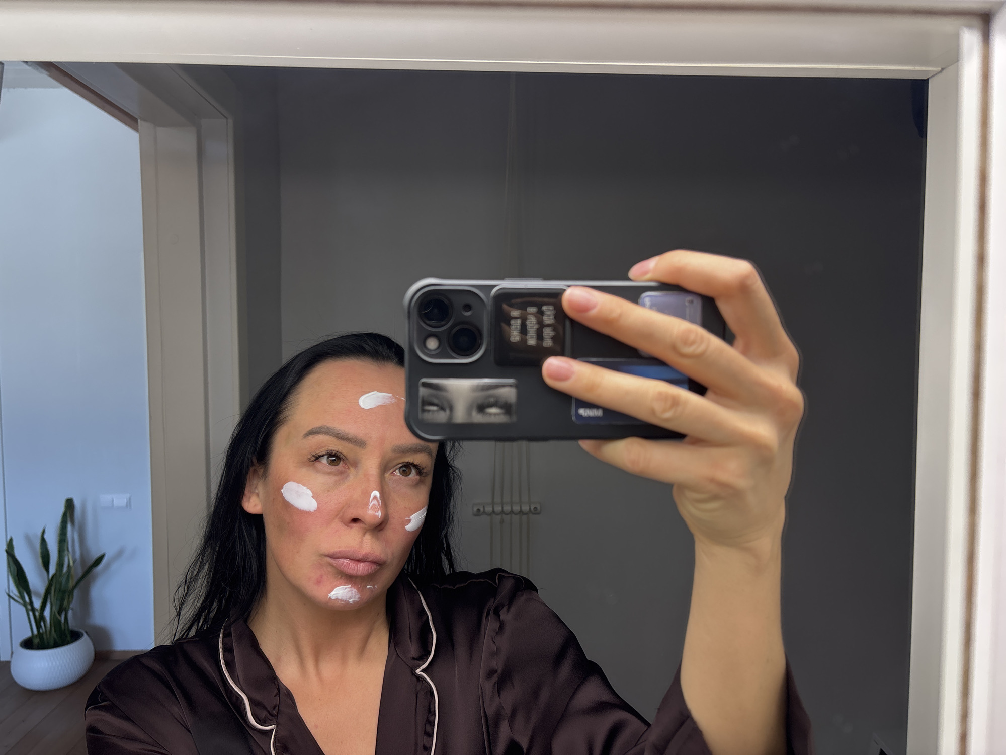 A woman stands in a bathroom, taking a selfie in the mirror. She has dark hair and is wearing a dark brown pajama top with a white trim. Her face has several spots of white cream on it. She is holding a smartphone in her right hand, which has a dark phone case with a graphic of eyes on it. The background shows a bathroom with a white framed mirror, and part of a doorway. The location is Chaikovsky, Russia.