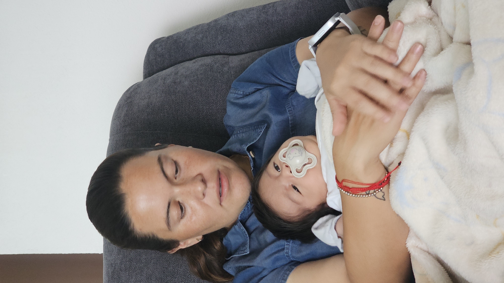 A person with dark hair and a denim shirt is lying on a gray couch, cradling a baby. The baby has dark hair and a pacifier in its mouth. The person is holding a white blanket. The person is wearing a silver smart watch on their left wrist and a red string bracelet on their right wrist. The location is San Bernardino Tlaxcalancingo, Mexico.