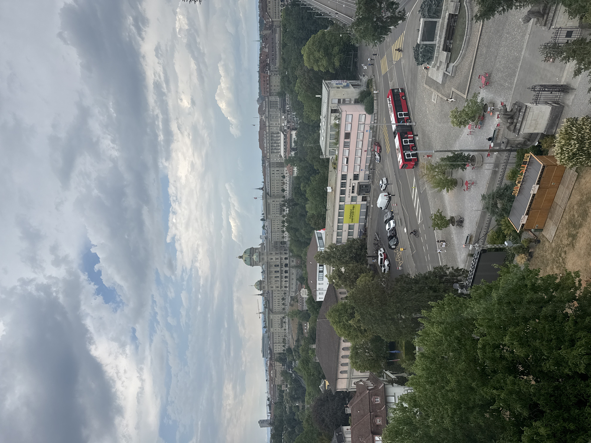 This is an overhead, wide-angle shot of a city street and buildings, captured on a cloudy day. The image is oriented vertically, with the top of the frame showing a sky filled with dramatic, grey clouds, hinting at potential rain or a storm.
The scene depicts a bustling urban environment. A bright red double-decker bus is a prominent object, driving on the paved road. Several cars are also visible on the street, some parked and others in motion. A few people can be seen walking on the sidewalks and crossing the street, some of whom are obscured by the angle.
In the background, a cityscape unfolds with numerous buildings of varying architectural styles. Some appear to be older, grand structures with domed roofs, possibly government or historical buildings, while others are more modern, multi-story edifices. Lush green trees are scattered throughout the scene, interspersed with buildings and lining some of the streets, suggesting a city with ample green spaces.
One building, colored pink, has "PH Bern" written on its side. Another building has yellow signage with text that is partially legible, appearing to be "Grünland ...".
The overall atmosphere is that of a normal day in a European city, with the overcast sky suggesting either early morning, late afternoon, or a generally cloudy day. No specific event seems to be taking place, but the presence of the bus and cars indicates ongoing daily activity.