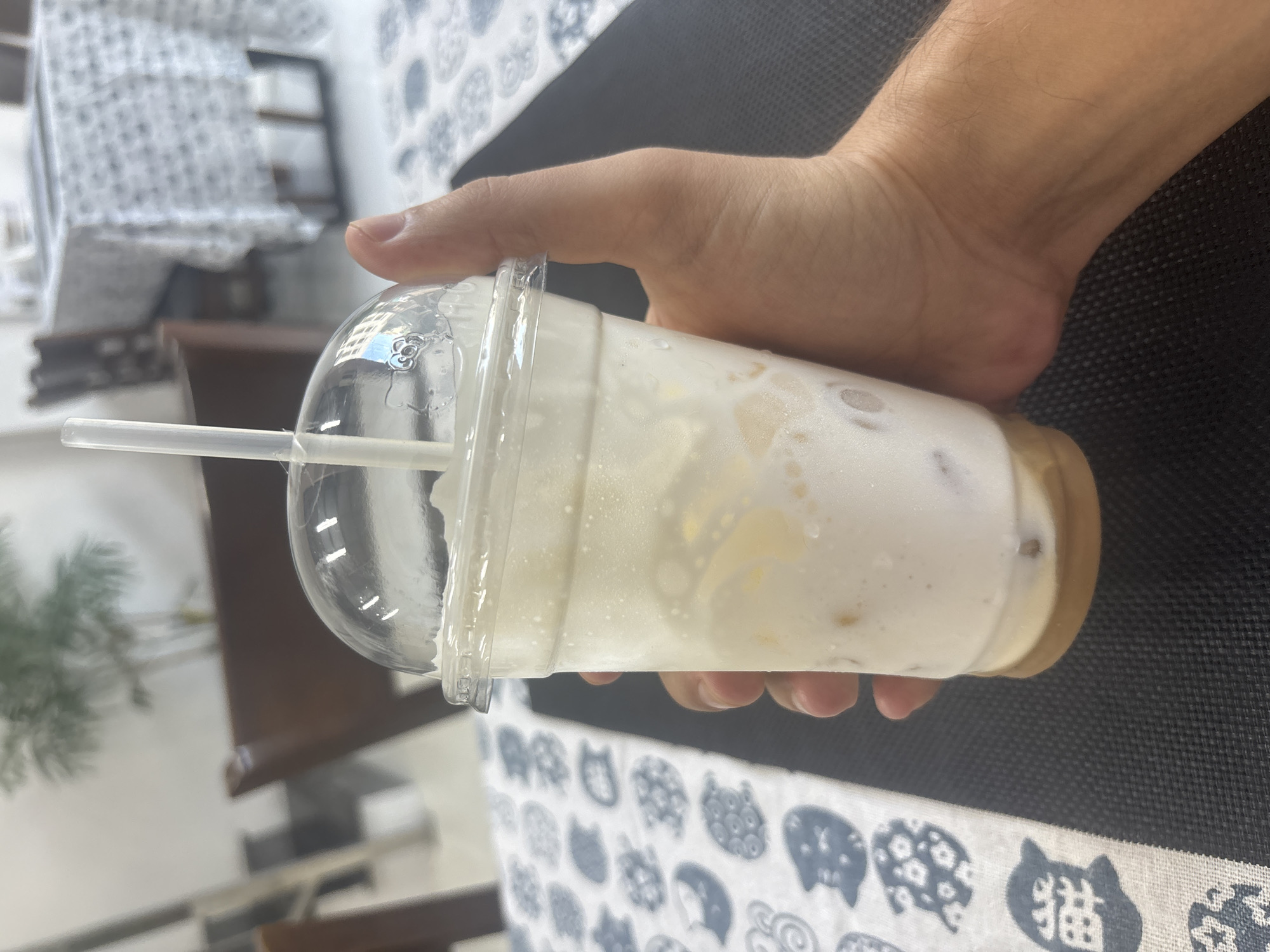 A close-up, slightly angled shot captures a hand holding a clear plastic cup filled with a frothy, light-colored beverage, likely a milkshake or smoothie, with a white straw inserted. The cup has a domed lid, and condensation is visible on its surface. The drink appears to have some solid pieces or ice at the bottom. The background suggests an indoor setting, possibly a cafe or restaurant, with blurred tables and chairs, and a patterned tablecloth. The lighting is bright, casting some reflections on the cup lid. The focus is on the drink and the hand holding it, with the background out of focus.