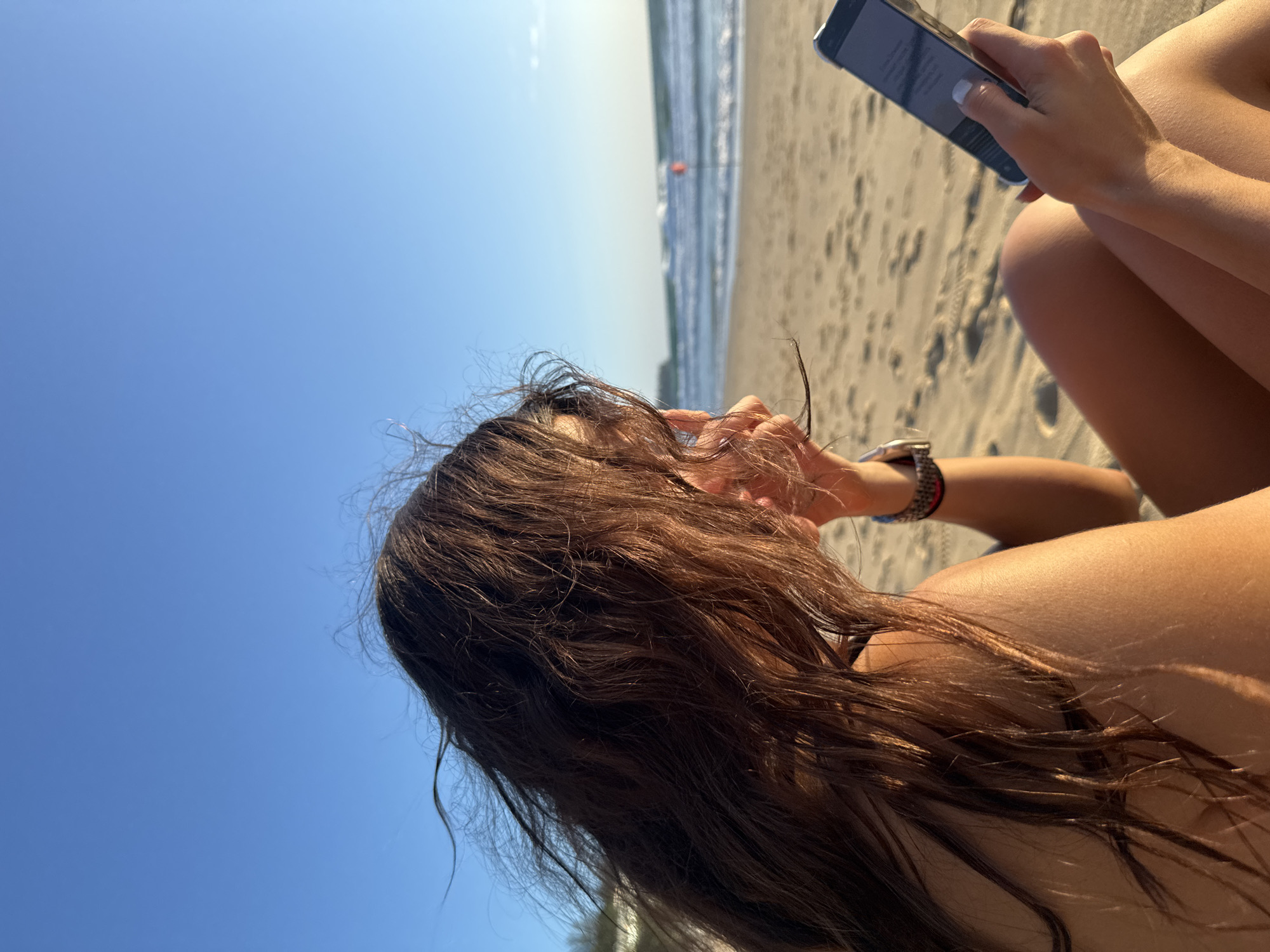 A woman with long, dark, windblown hair is sitting on a sandy beach in Choeng Thale, Thailand, looking at her smartphone. The sun is shining, casting shadows on the sand and highlighting the woman's tanned skin. The ocean is visible in the background with gentle waves rolling towards the shore. The sky is clear and blue.