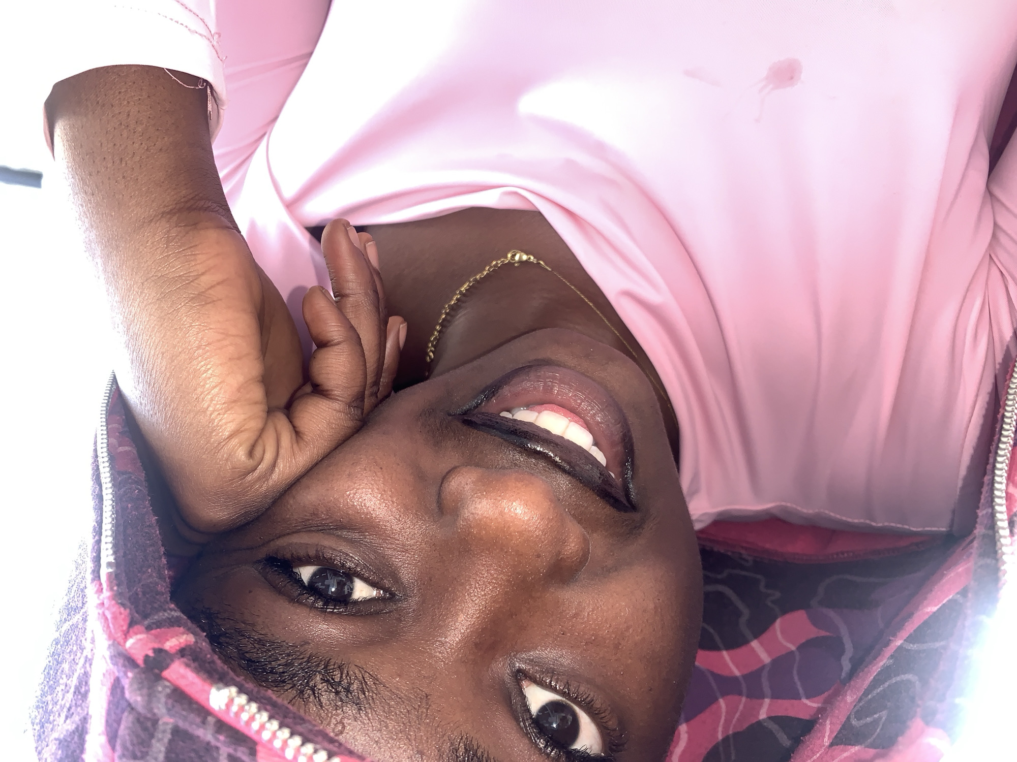 A close-up, angled shot shows a young Black woman with her head tilted and resting on her hand, her right cheek pressed against her palm. She is wearing a pink hooded jacket and a pink shirt. The jacket is unzipped, revealing the pink shirt underneath. Her dark hair is visible under the hood, and she has on dark eyeliner and lip gloss. A thin gold chain is visible around her neck. The background is out of focus and appears to be an indoor setting. There are no visible landmarks or indicators that would suggest the location is Gura Top, Nigeria.