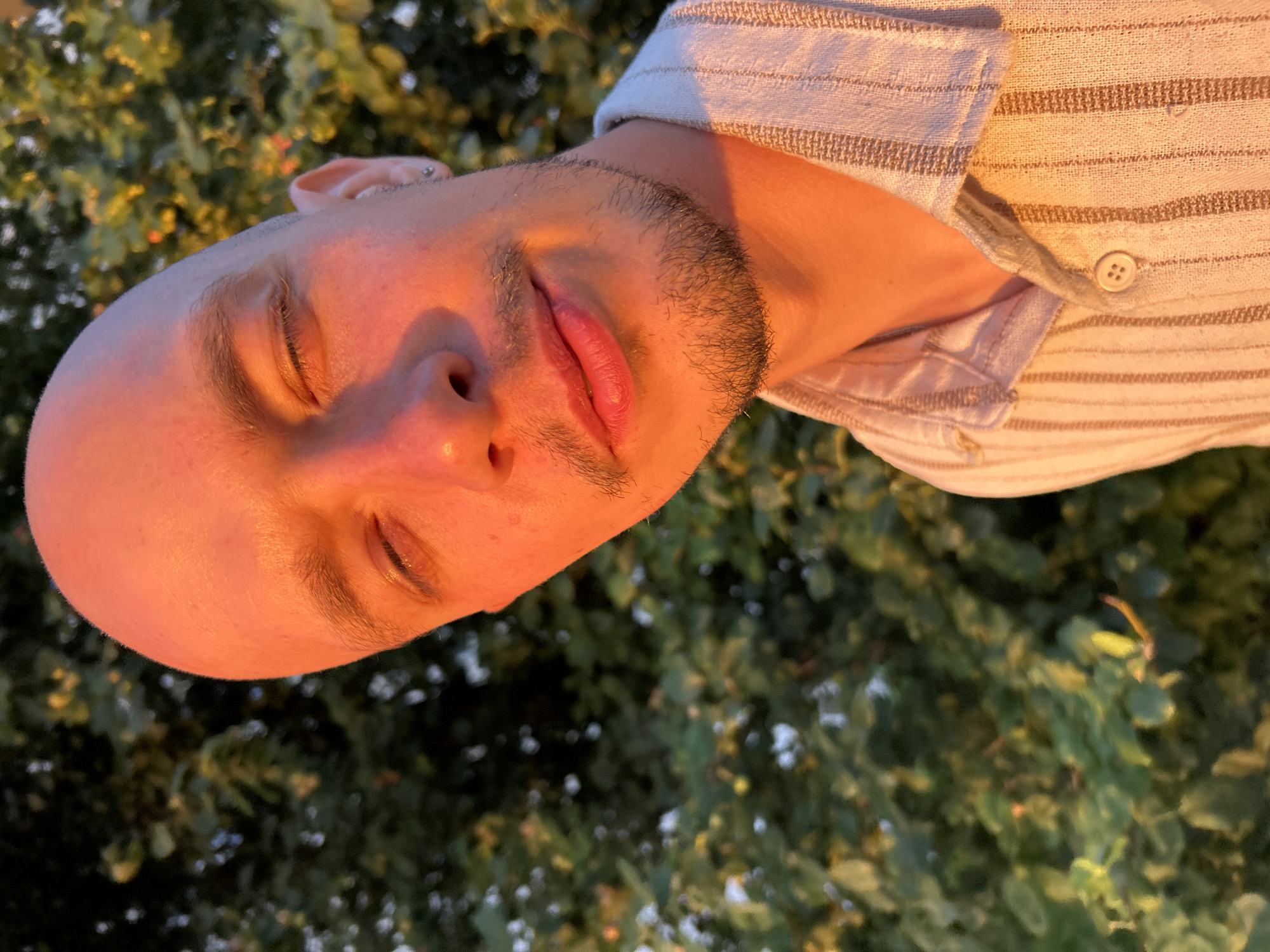 A close-up, slightly angled, eye-level shot shows a bald individual with closed eyes and a faint smile, bathed in warm, golden-hour light. The person is wearing a light-colored, striped shirt with a collar. The background is filled with blurred green foliage, likely trees, suggesting an outdoor setting. The lighting suggests it is either sunrise or sunset. There are no other people or significant objects visible. The image does not provide any specific indicators of its location within the city of Chaikovsky, Russia.