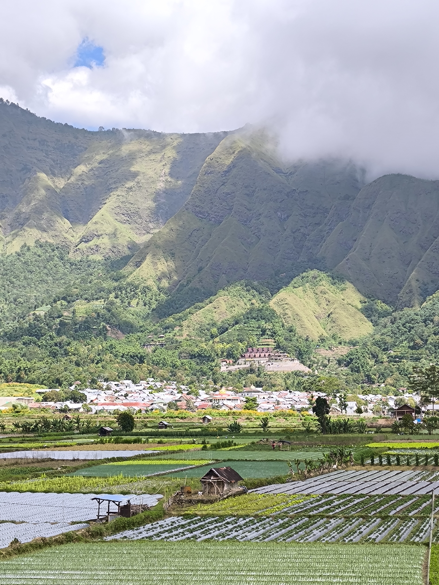 The image shows a wide-angle, daytime view of the Sembalun Bumbung area in Indonesia. In the foreground are agricultural fields with rows of crops, some covered in plastic sheeting. A small wooden shack and a structure with a blue roof are visible among the fields. A village is situated in the midground, with white houses and red roofs. Behind the village, a steep, green mountain range rises, with the peaks partially obscured by clouds. The sky is overcast with some patches of blue.
