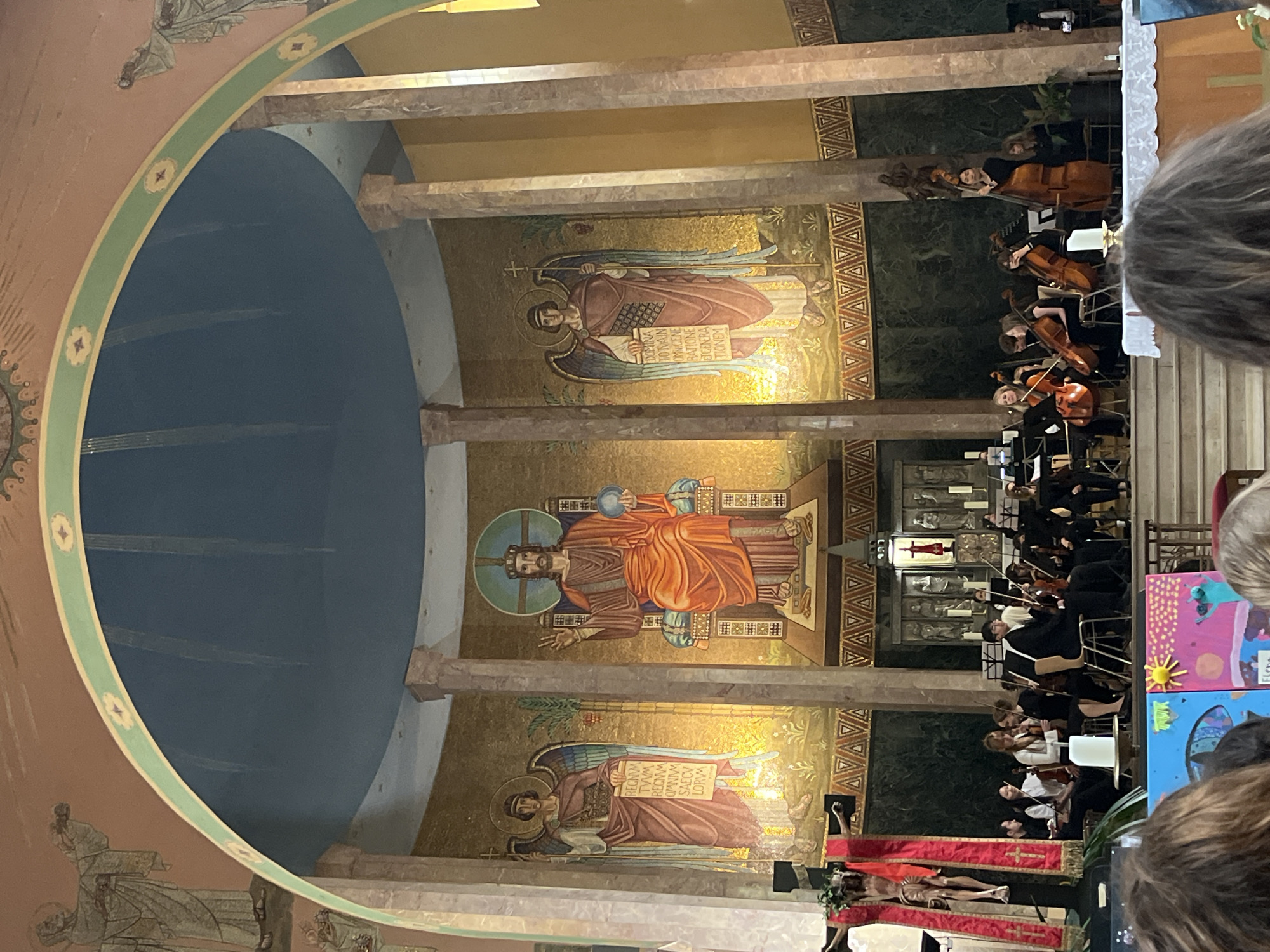 Here is a detailed description of the image:
**Scene:** The image captures the interior of a church, likely in Saarbrücken, Germany, given the presence of Byzantine-style mosaic artwork on the walls, which is characteristic of the St. Michael Basilica located there. An orchestra performs in the area in front of the apse. There is a crucifix on the right side of the orchestra. The scene is viewed from the audience perspective.
**Artwork:** The walls of the apse are adorned with mosaics of Byzantine style. The central mosaic appears to depict Christ Enthroned in Majesty, as a full figure, with the world in his left hand. Above and below this figure are two winged figures with writing on the tablets, in an illuminated gold background.
**People:** The foreground contains audience members; hair visible in the frame of people sitting or standing in front of the picture taker. The church setting suggests a potentially religious gathering or concert event.
**Environment:** The location is a church, recognizable by the architectural design of the apse. A dome is visible over the apse. The lighting appears to be a mixture of natural and artificial light.
**Details and Objects:**
* **Mosaic artwork:** Large, Byzantine-style mosaics depicting religious figures.
* **Crucifix:** A crucifix is to the right of the orchestra
* **Music Equipment:** Instruments, music stands, and related items associated with the orchestra.
* **Audience:** The audience members are visible in the lower and side regions of the frame.