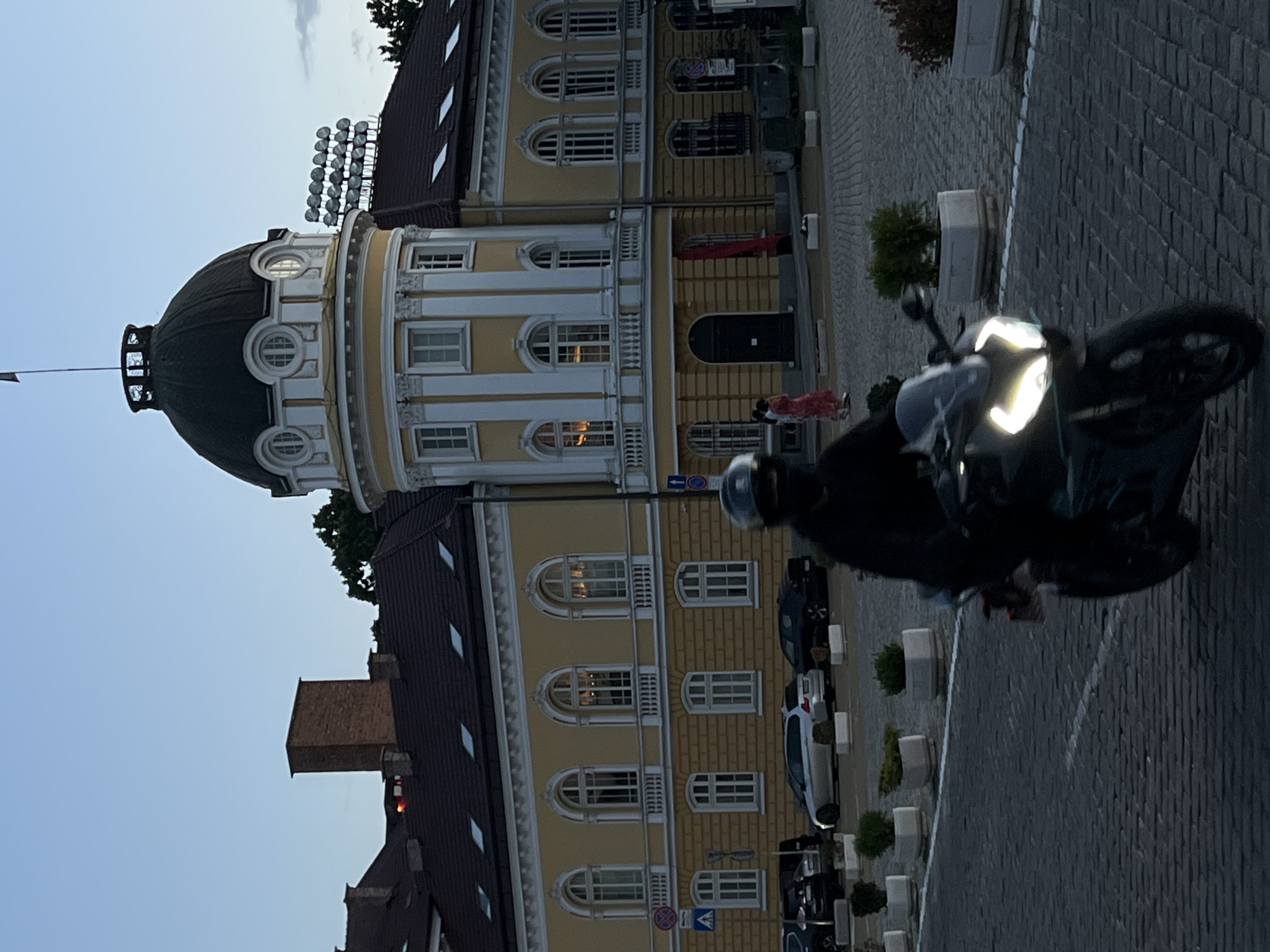 An eye-level, medium shot captures a motorcyclist in motion from the right side of the frame to the left, on a cobblestone street in front of a large, yellow building with a domed structure. The motorcyclist is wearing a black helmet and dark clothing. The building features a prominent dark dome with decorative white trim and arched windows. The facade of the building is yellow with white architectural details and multiple arched windows. Two figures, a woman in a red dress and another person, are visible walking in front of the building. Cars are parked on the left side of the image. Planters with greenery are placed along the street. The sky is a pale blue, indicating it is either dawn or dusk. The scene appears to be located in the Largo area of Sofia, Bulgaria, specifically in front of the former Party House, now housing government offices and the National Art Gallery.