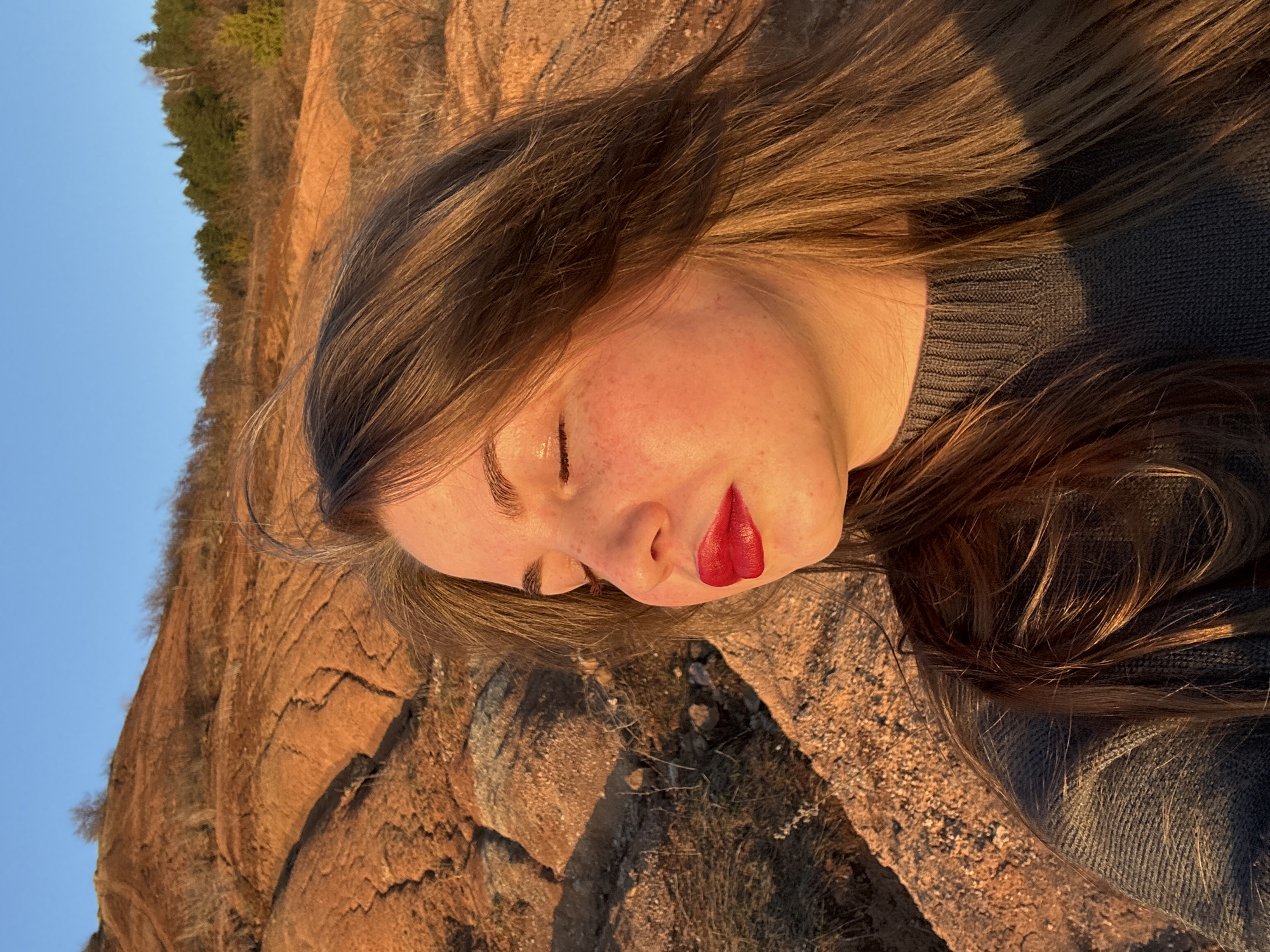 A young woman with fair skin, freckles, and long brown hair lies on her side on a rocky surface in Ольховка, Russia. She wears a dark grey knit sweater and red lipstick. A rocky hill with sparse vegetation and a blue sky are visible in the background. The scene is lit by warm, golden light, suggesting it is near sunset or sunrise.