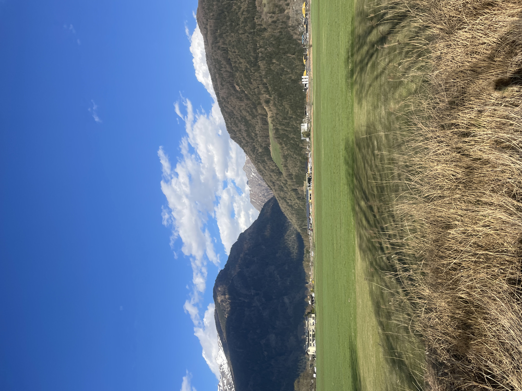 The image captures a landscape in Sterzing-Vipiteno, Italy, oriented vertically. The sky is blue with scattered white clouds. Adjacent to the sky is a mountain range. The lower slopes of the mountain lead to a green field, separated from dry, brown vegetation in the foreground. Buildings are situated at the base of the mountain. Tree shadows are visible on the green field.