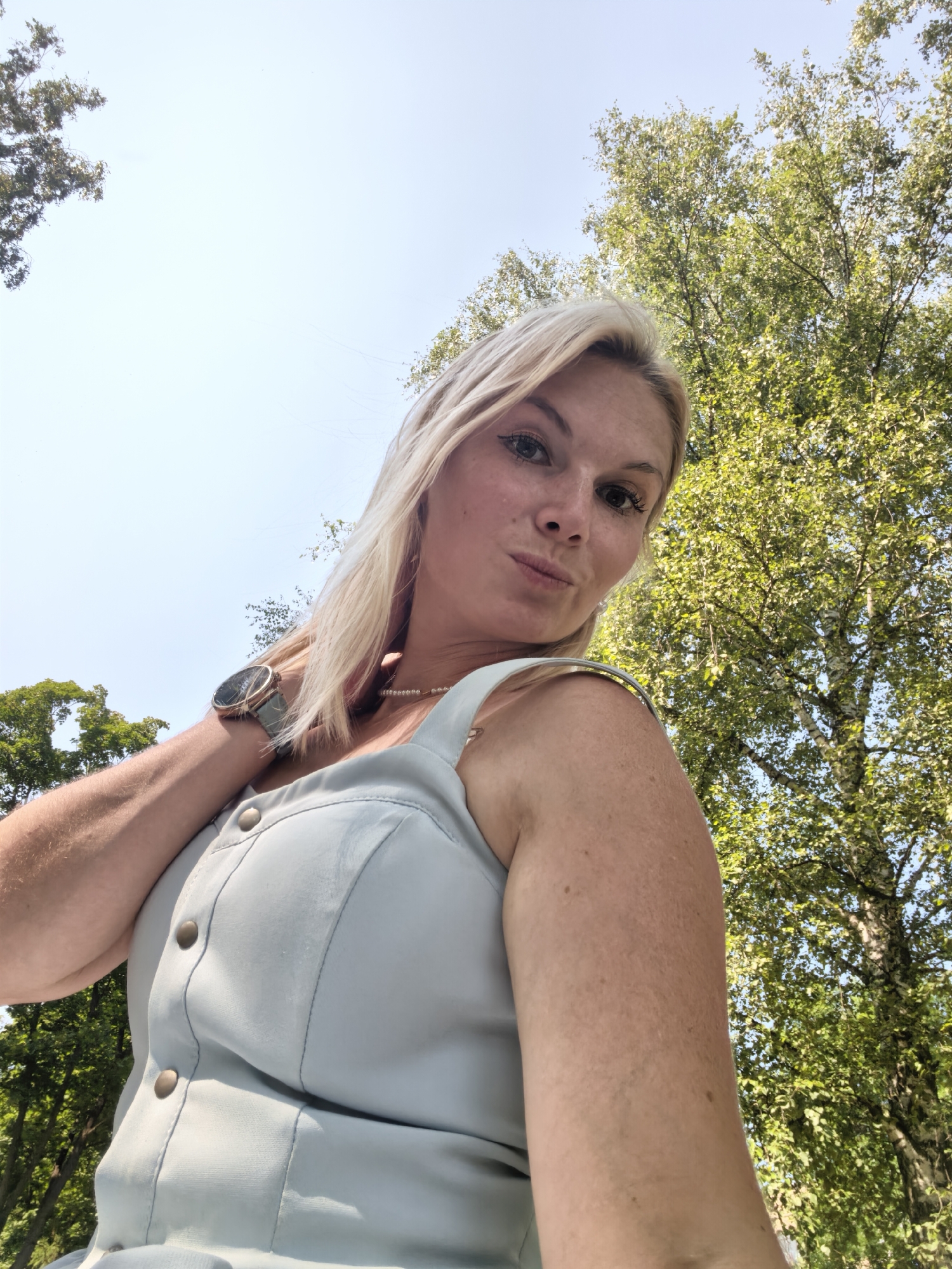 An outdoor, low-angle, eye-level, waist-up, close-up selfie of a blonde woman. The woman is facing the camera with her head tilted to the left, lips puckered, and her right hand resting on her neck. She is wearing a light blue button-up dress and a watch on her left wrist. The background consists of a bright, pale sky and lush green trees. The image does not contain any information regarding the location in the city of Sterlitamak, Russia.
