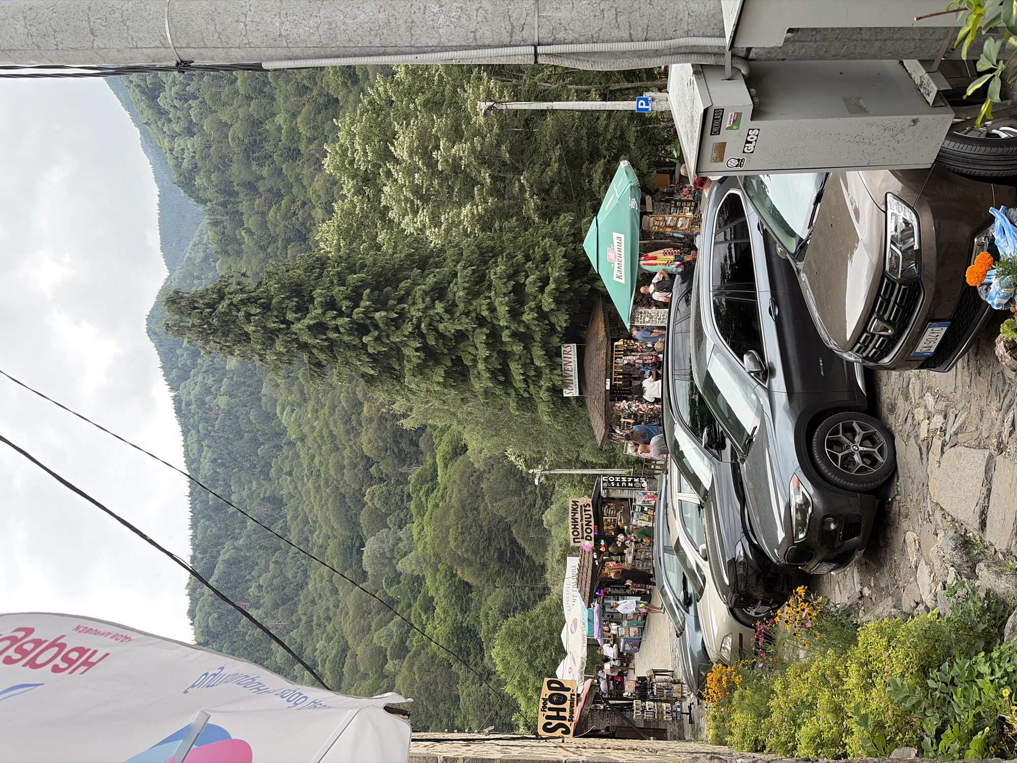 The image depicts a street scene in Rila Monastery, Bulgaria, characterized by a mountainous backdrop covered in lush green forests under a cloudy sky. In the foreground, a line of parked cars, including a brown Dacia Logan and a dark gray BMW, are parked along a cobblestone street. Adjacent to the street, a row of souvenir shops and food stalls are set up, with awnings and umbrellas providing shade. People can be seen browsing the shops and interacting with vendors. One shop displays a sign for "Food - Shop - Souvenirs." Another stall features "ПОНИЧКИ DONUTS" (Donuts in Bulgarian). A large green umbrella with the text "Каменица" (Kamenitsa, a Bulgarian beer brand) is visible, possibly indicating a refreshment stand. A concrete utility box with stickers is positioned on the right side of the image. The overall atmosphere suggests a bustling tourist area within a natural setting.