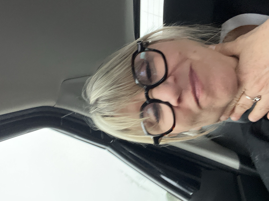 The image displays a female individual situated within the interior of a vehicle, in what is identified as Vidauban, France. The subject, a Caucasian woman with light blonde hair styled with bangs, is wearing dark-rimmed, round-shaped eyeglasses with a tortoiseshell pattern. Her face is directed towards the viewer, with her lips forming a slight upward curve. Her right hand is positioned against the right side of her neck, with the thumb near the jawline and fingers extending downwards. A silver-colored ring is visible on the fourth finger of her right hand. She is dressed in a dark garment, possibly a jacket or shirt, over a visible light-colored collar. The vehicle's interior is visible behind her, including a dark A-pillar and a light grey headliner. Through the front left window, an indistinct, bright, light grey outdoor environment, potentially an overcast sky, is visible.