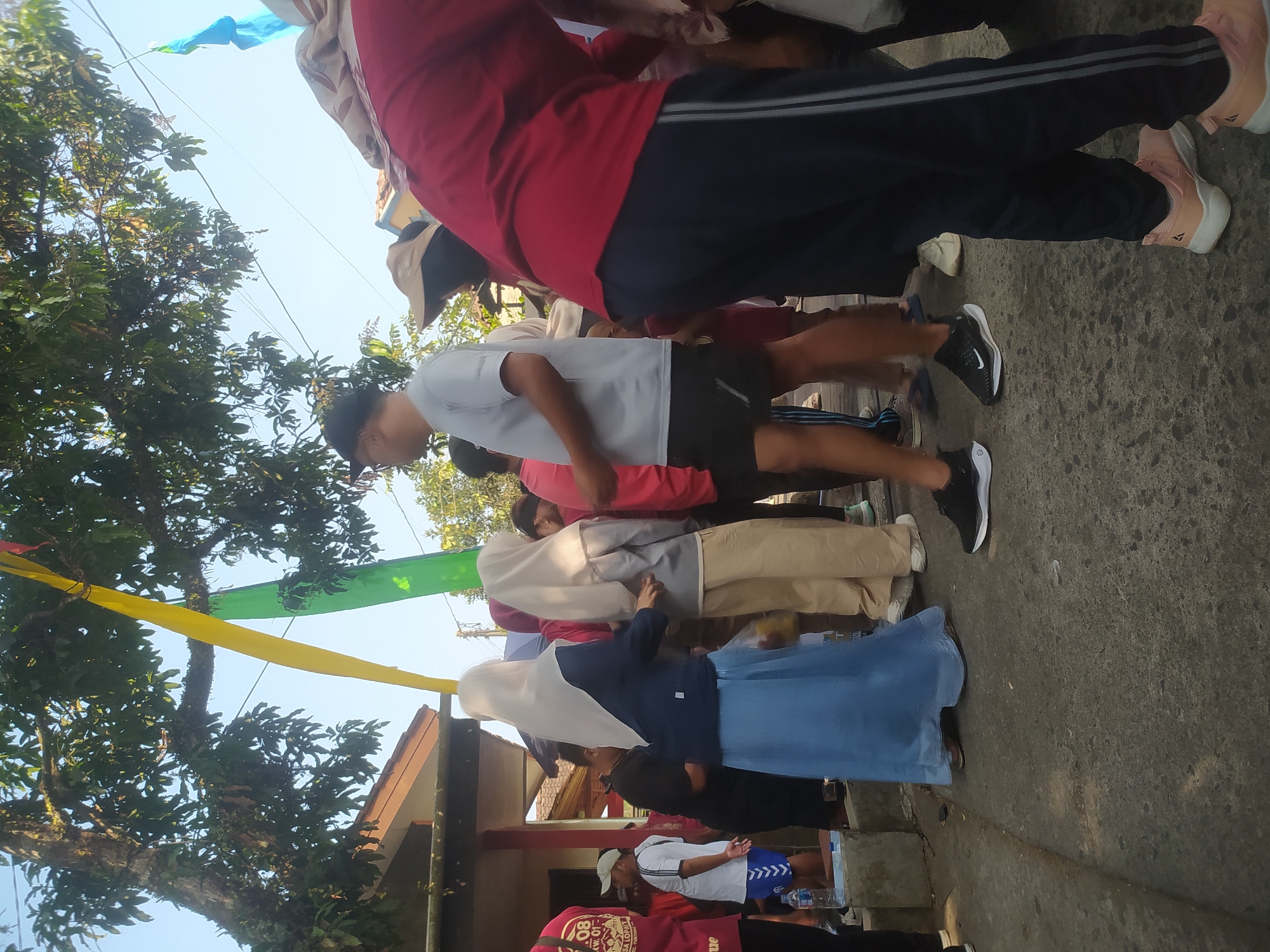 The image depicts an outdoor scene in the city of Lopait, Indonesia, likely during an event or gathering. The foreground is dominated by the legs and lower torsos of several individuals, suggesting a crowd. A person in a red long-sleeved shirt and dark pants with stripes down the side is prominently positioned in the right foreground, their body angled away from the viewer. To the left, a man in a light gray t-shirt and black shorts is facing left in profile. Further left, a woman in a long blue skirt and a light-colored headscarf is interacting with another person partially obscured. Another woman, also in a light-colored headscarf, stands next to her, wearing beige pants. In the background, a building with a brown roof is visible, and trees with green foliage are present. Colorful streamers, including yellow and green, are strung across the scene, possibly indicating a celebratory or organized event. The ground appears to be paved. The overall atmosphere suggests a lively community gathering.