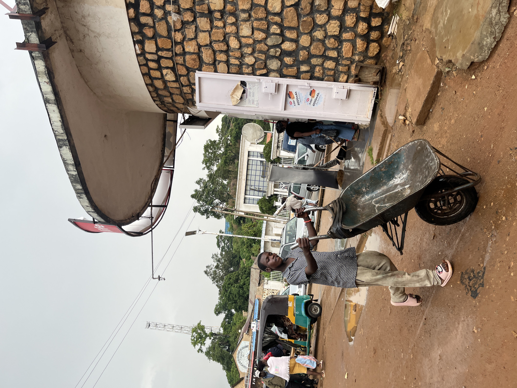In Jos, Nigeria, a young boy is pushing a wheelbarrow down a wet, muddy street. The boy is wearing a black and white patterned shirt and grey pants, with pink slides on his feet. Behind him, other people and vehicles, including an auto-rickshaw, are visible, suggesting a busy street scene. The building in the background has a distinctive curved roof and is constructed with stone walls. The sky is overcast, indicating a cloudy day.