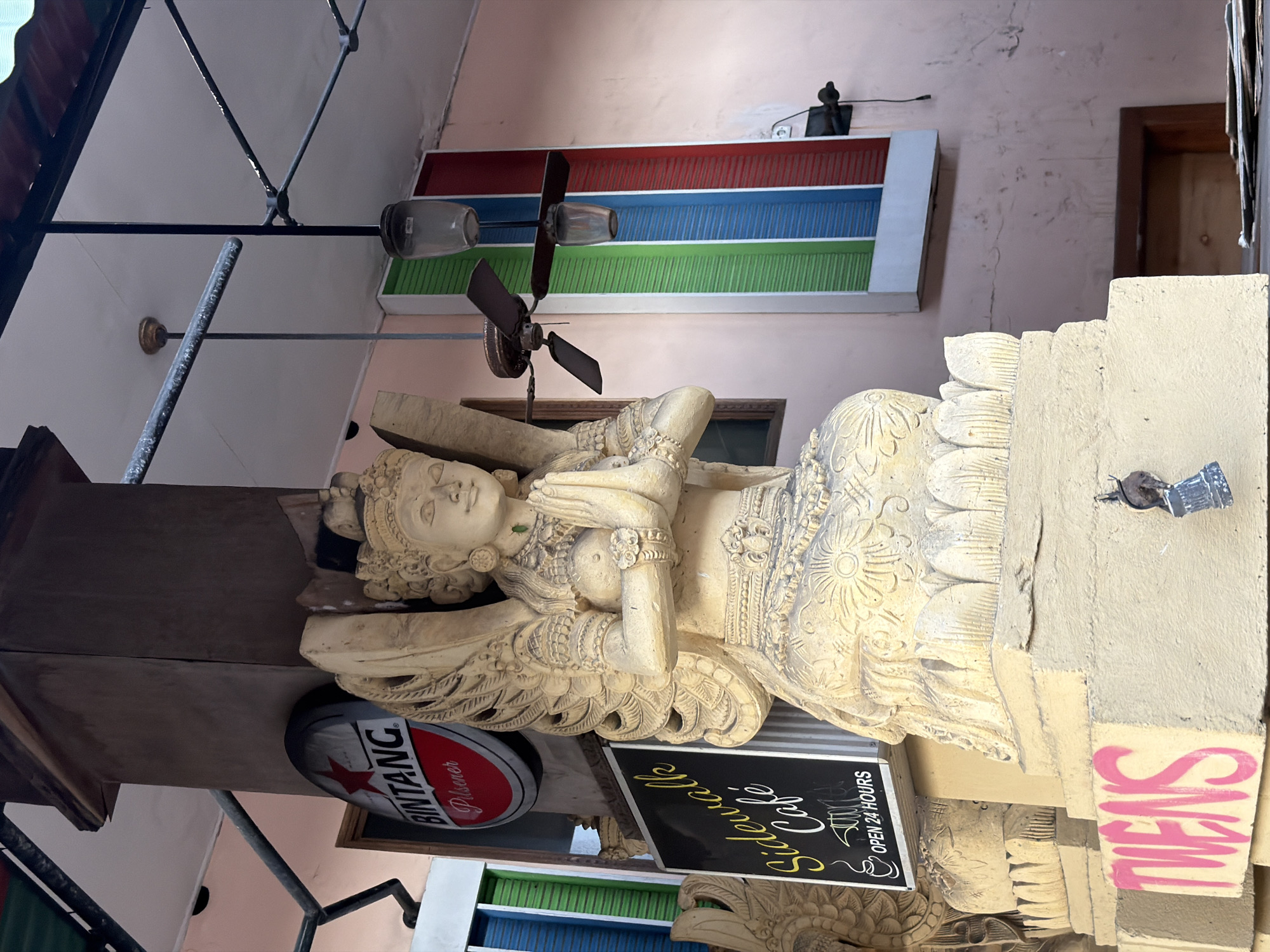 The image captures a statue of a winged female figure in a prayer pose, situated in front of a building with a pink wall and a striped facade of green, blue, and red. A "Bintang Pilsener" sign is visible to the left of the statue, and a sign for "Sidewalk Café OPEN 24 HOURS" is positioned below it. Two ceiling fans hang from the ceiling above, one of which is actively spinning. The statue rests on a pedestal that has "TWENS" spray-painted in pink on its side. The overall scene suggests a street-level view of a cafe or establishment in Kuta, Indonesia, likely a tourist area given the presence of the Bintang beer advertisement and the cafe sign.