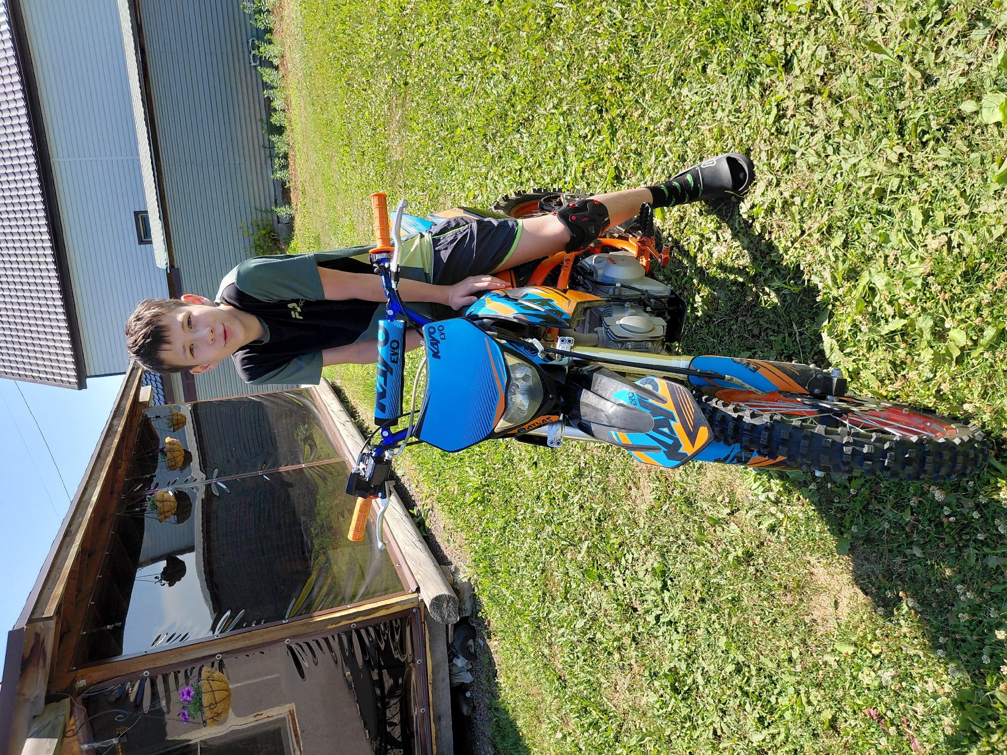 A boy sits on a dirt bike outdoors on a sunny day. He is wearing a black and green t-shirt, black shorts, black knee pads, and black socks with green accents. The dirt bike is blue and orange with the brand "KAYO" visible on the front. The background shows a wooden structure with hanging flower baskets and a light blue house with a tiled roof. The ground is covered in green grass. The scene appears to be in a residential area, possibly a dacha or country house outside of Novosibirsk, Russia.