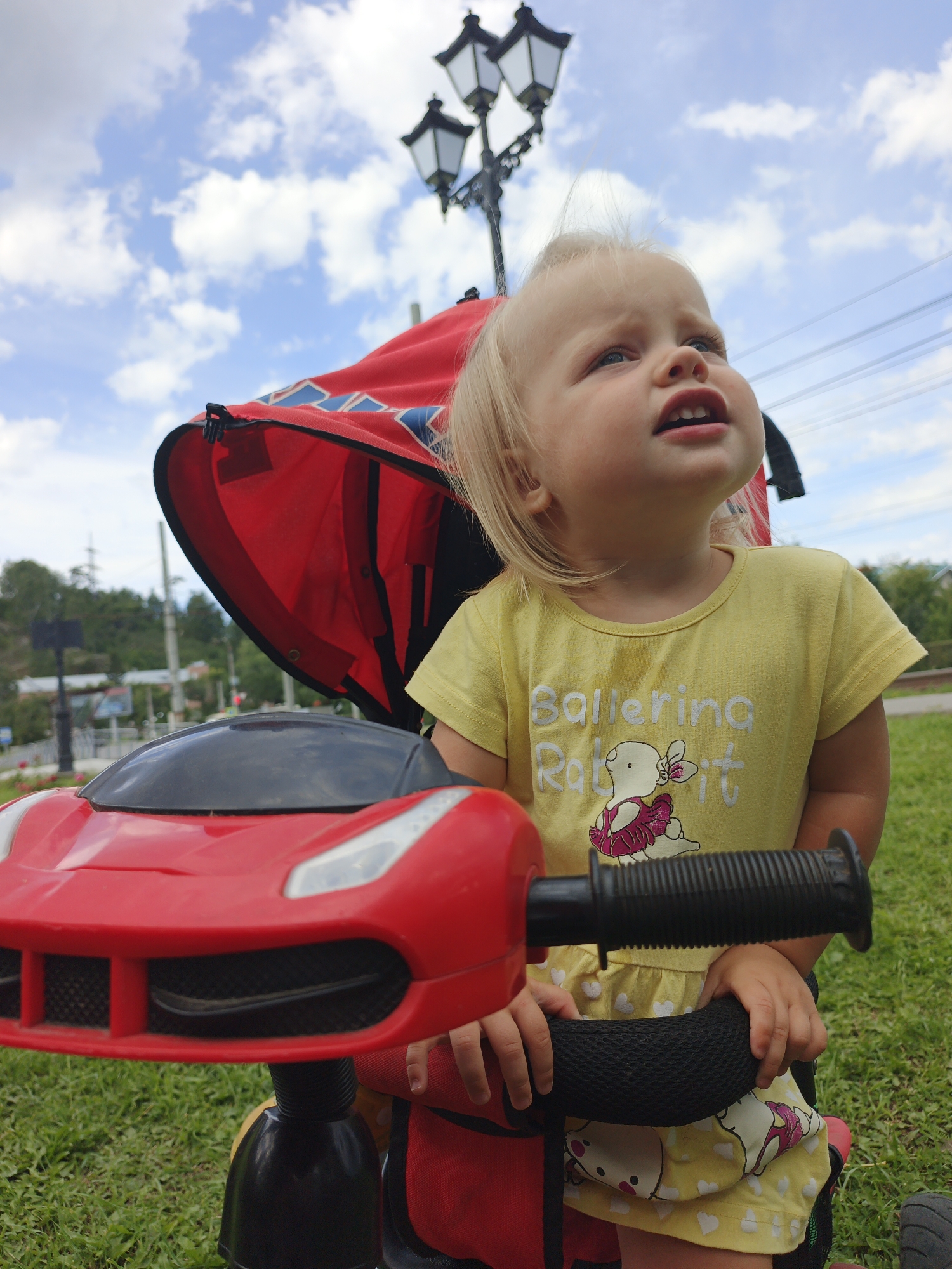 A young Caucasian female with blonde hair and blue eyes, wearing a yellow t-shirt with a "Ballerina Rabbit" graphic and a yellow dress with white hearts, sits in a red ride-on toy car outdoors. She is looking up and to the right. The toy car has a black steering wheel. In the background, a red canopy from a stroller is visible, along with a lamppost with three lights, trees, and a cloudy sky. The scene appears to be in a park or outdoor public space.