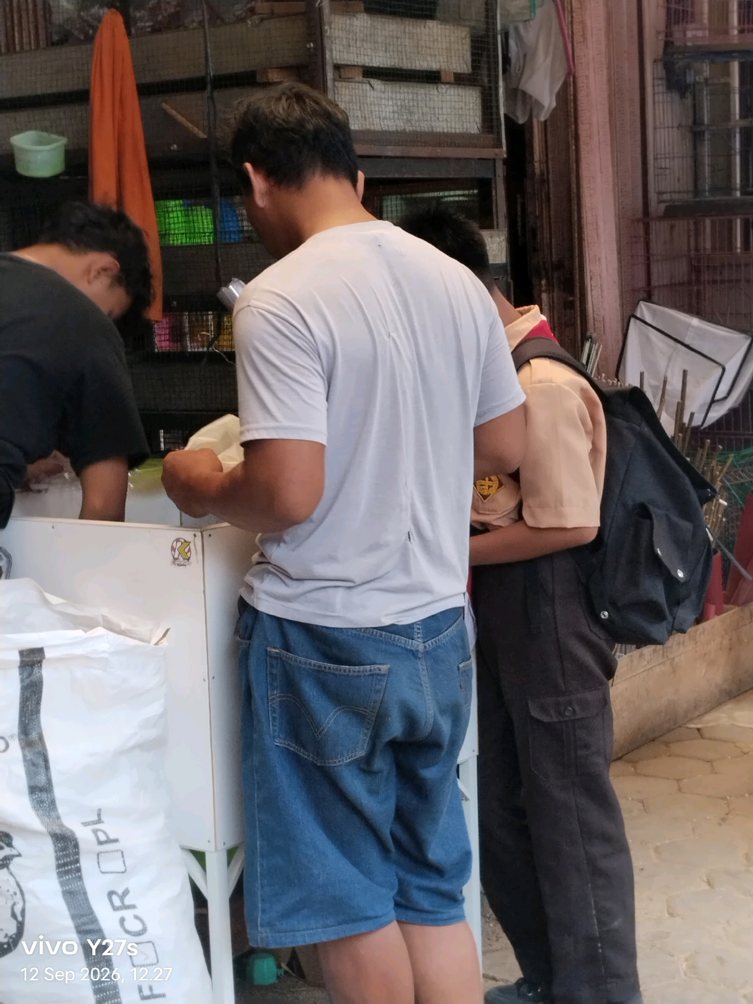 Two individuals are visible in the foreground, with the primary focus on a person wearing a light grey t-shirt and blue denim shorts. This person is facing away from the camera, holding a white object, possibly a bag of goods, in their right hand. To their left, another person, partially visible and facing away, is wearing a black t-shirt. Behind these individuals, a person in a tan uniform, possibly a scout uniform, with a dark backpack is also present. The background shows shelves filled with items, cages, and what appears to be an outdoor market or shop setting, with a concrete floor and a wall. The scene is set in Trimulyo, Indonesia, as indicated by the metadata.
